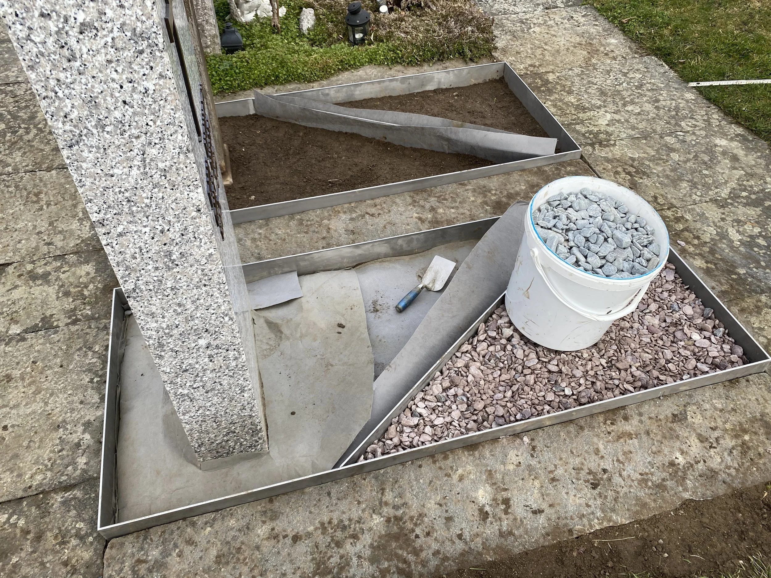 Grundrahmen für eine Gartenbeet mit Steinen, Erde und Bauteilen zur Umsetzung. Ein Eimer mit Kies und Werkzeuge sind sichtbar.