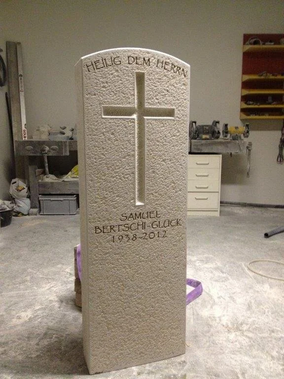 Granitgrabstein mit Kreuz und Inschrift, im Hintergrund Werkstattraum mit Werkzeugen