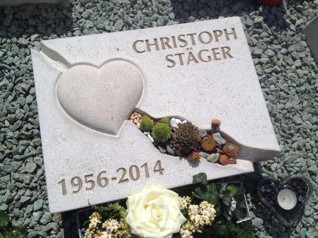 Grabstein mit Herzsymbol und Sukkulenten, mit Namen Christoph Stäger, geboren 1956 und gestorben 2014, liegend auf einem Kiesbett, mit Blumen und Kerzen am Grab.