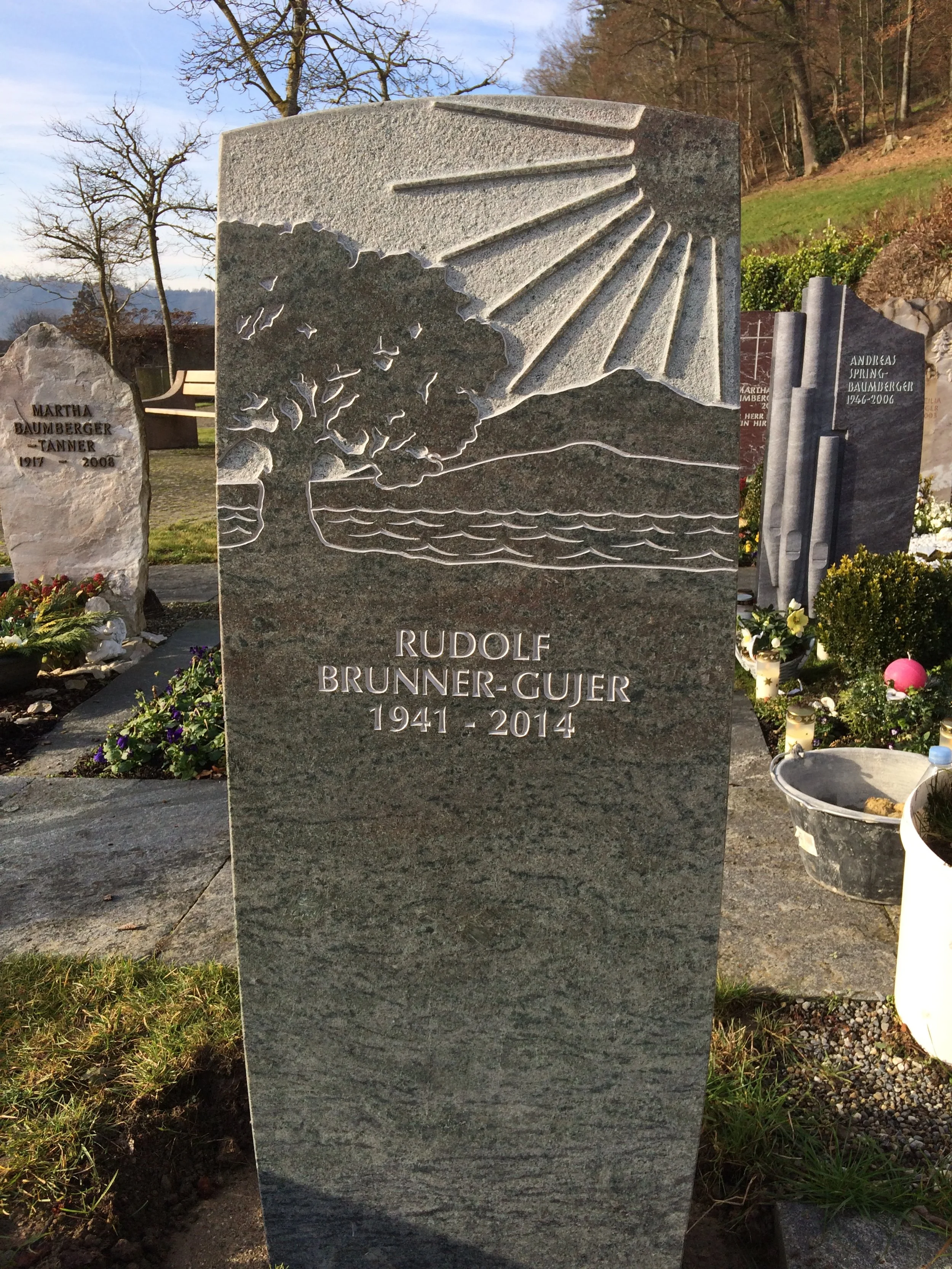 Ein Grabstein aus grauem Stein mit einer Gravur eines Sonnenstrahls, eines Baumes, Wasser und Hügeln, mit dem Namen Rudolf Brunner-Gujer (1941-2014)