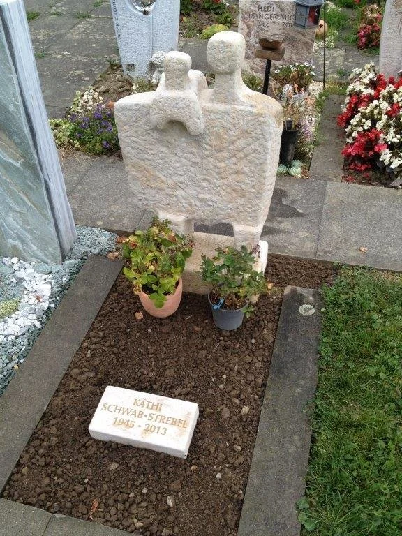 Ein Grabstein mit der Aufschrift »KÄTHI SCHWAB-STRAßEL 1948 - 2013« vor einem großen, abstrakten Steinkunstwerk auf einem Friedhof, umgeben von Blumen und Pflanzen.