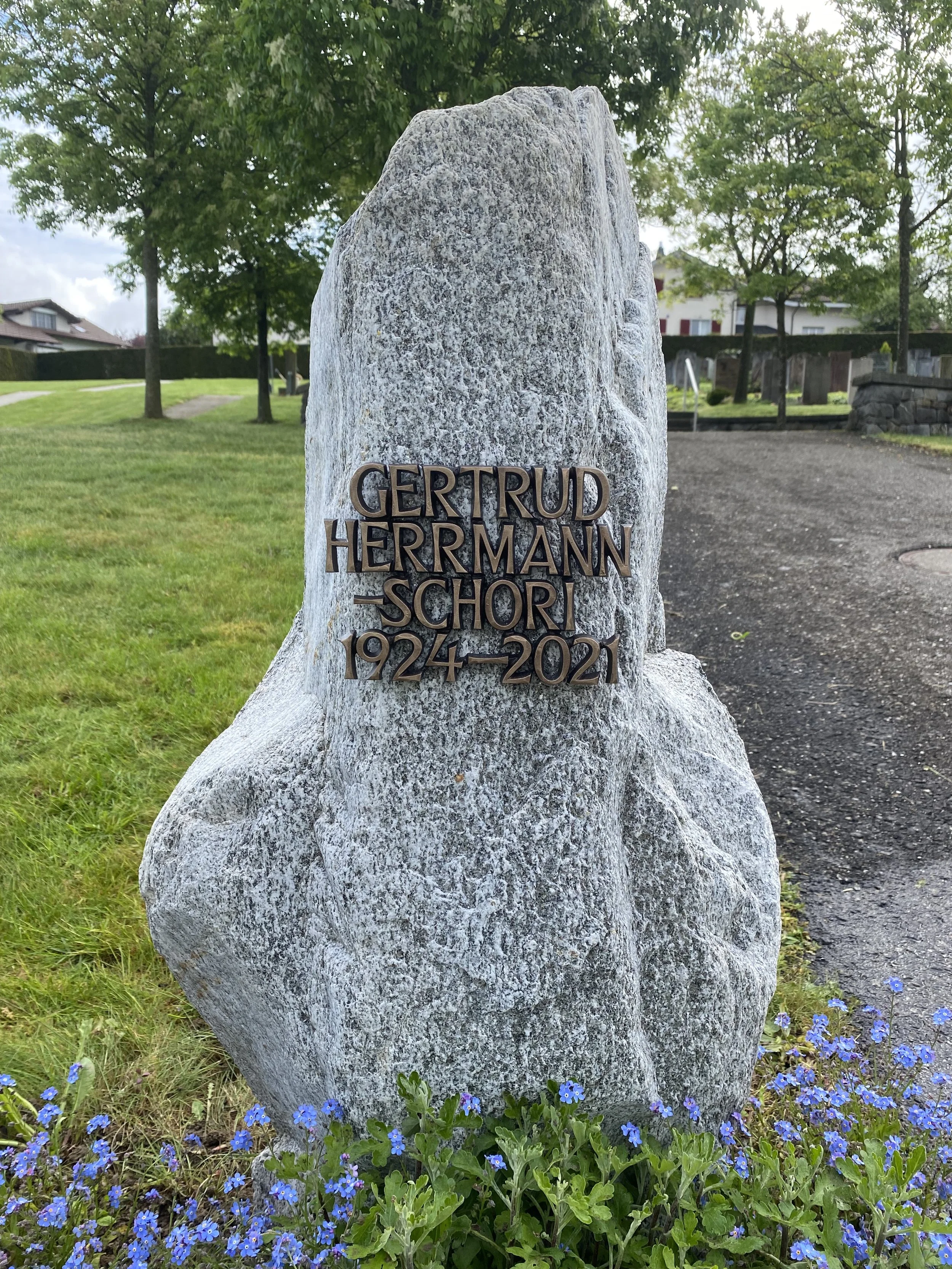 Grabstein mit Metallbeschriftung für Gertrud Herrmann-Schori, 1924-2021, in einem Garten mit grünem Rasen, Bäumen und blauen Blumen am Fuße des Steins.