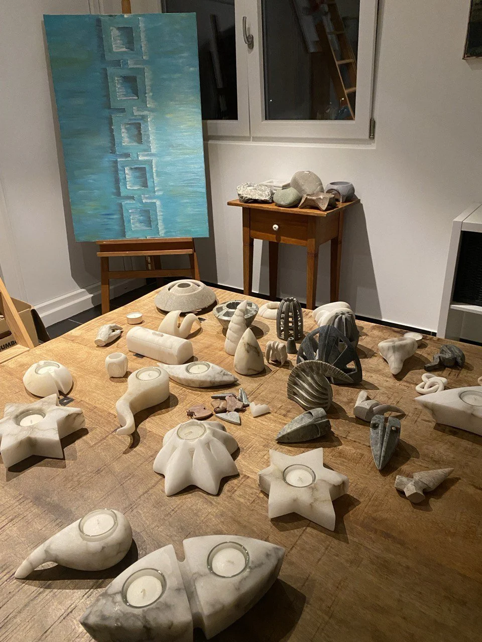 Tafel mit verschiedenen geschnitzten Kerzenhaltern und Skulpturen, im Hintergrund ein Tanzbild aus blauen Quadraten, an der Wand eine kleine Holzkommode mit Steinen darauf, eine Fenster