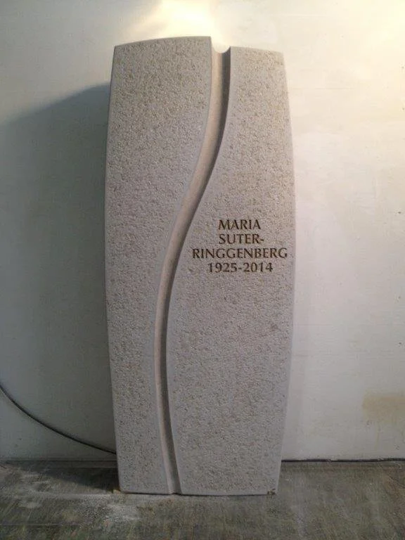 Grabstein mit eingraviertem Namen und Daten, aus hellem Stein mit geschwungener Linienführung.