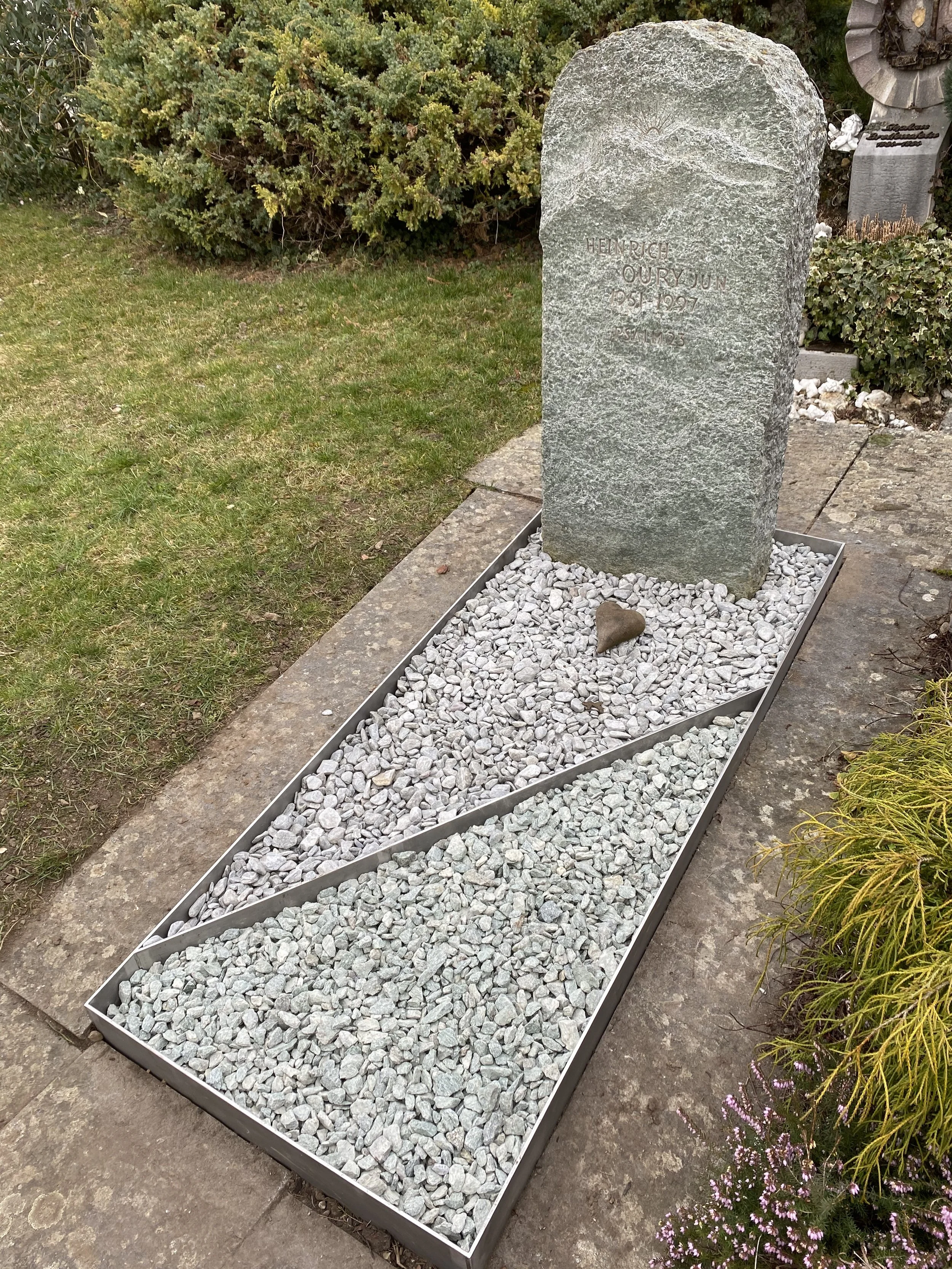 Ein Grabstein auf einem Begräbnisplatz, umgeben von kleinen weißen Steinen und einem Metallrahmen. Der Grabstein ist aus Stein, mit eingraviertem Text. Es gibt um den Bereich herum verschiedene Pflanzungen und Büsche.