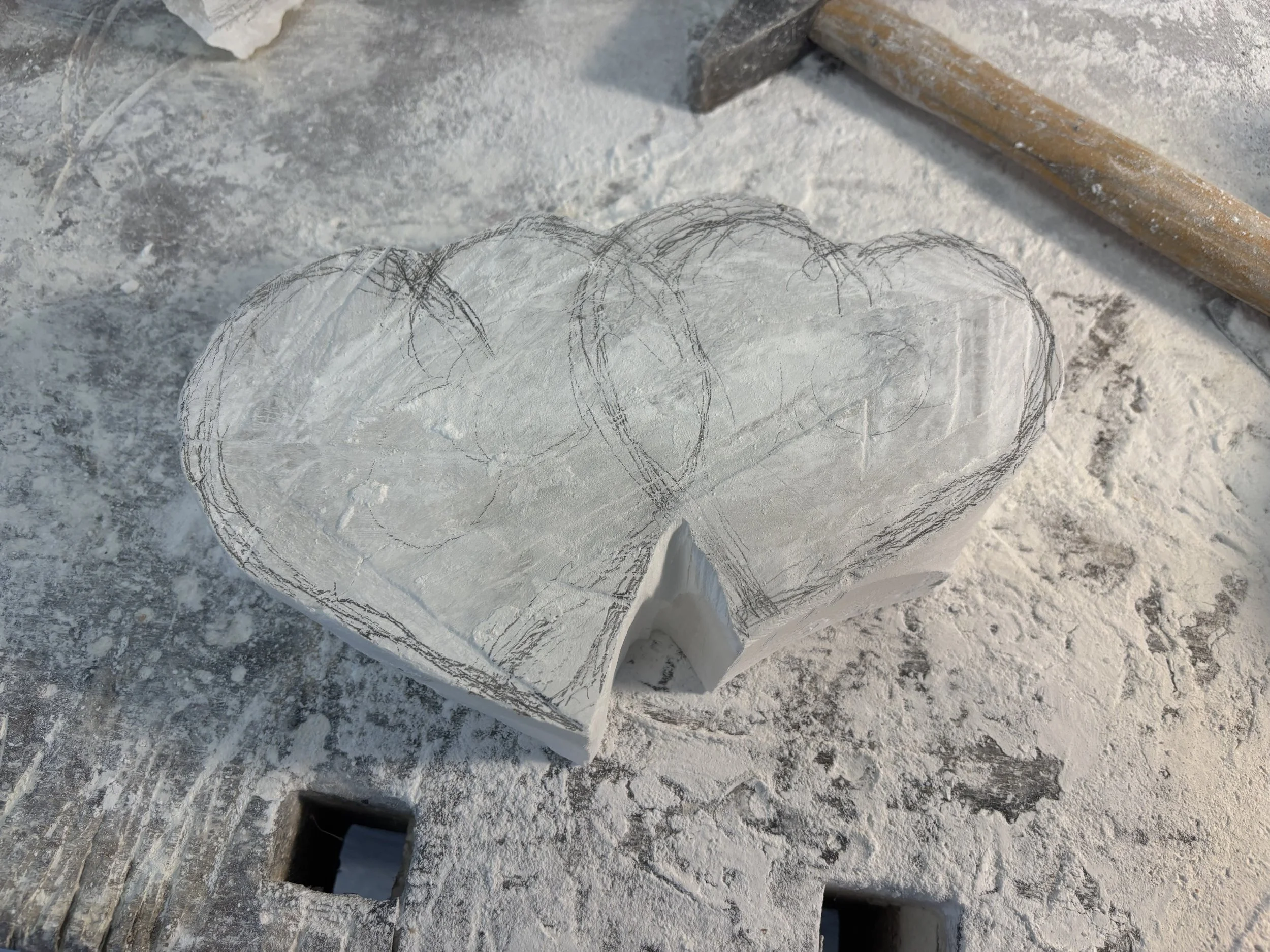 Holzblock in Form eines Herzens, umrandet mit Bleistiftlinien, auf einem verschmutzten Werkstück mit Staub, Werkzeug und Hammer im Hintergrund