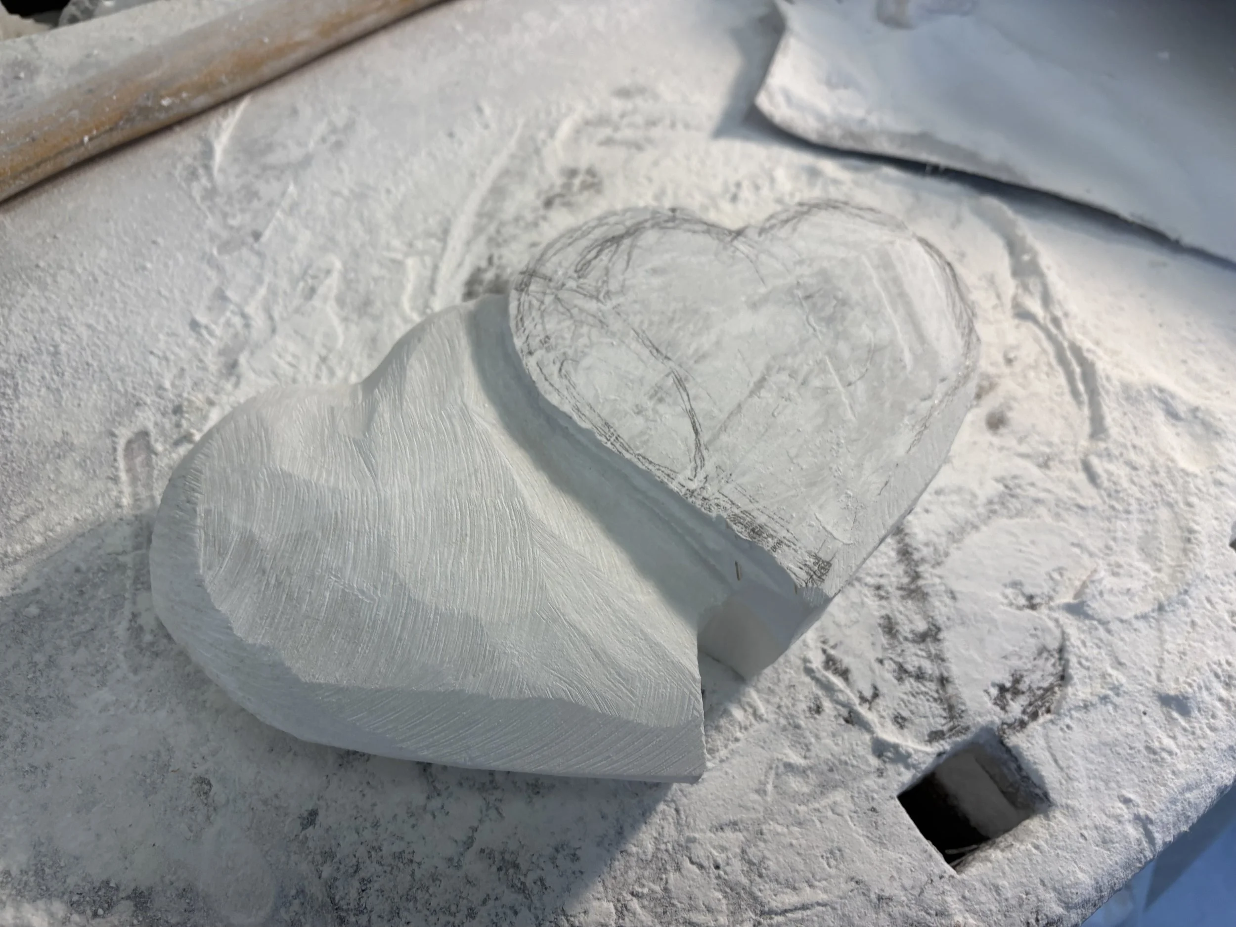 Ein Herz aus Holz wird in weißer Pulvererde geschnitzt, auf einer Arbeitsfläche mit Werkzeugen und einer Schablone.
