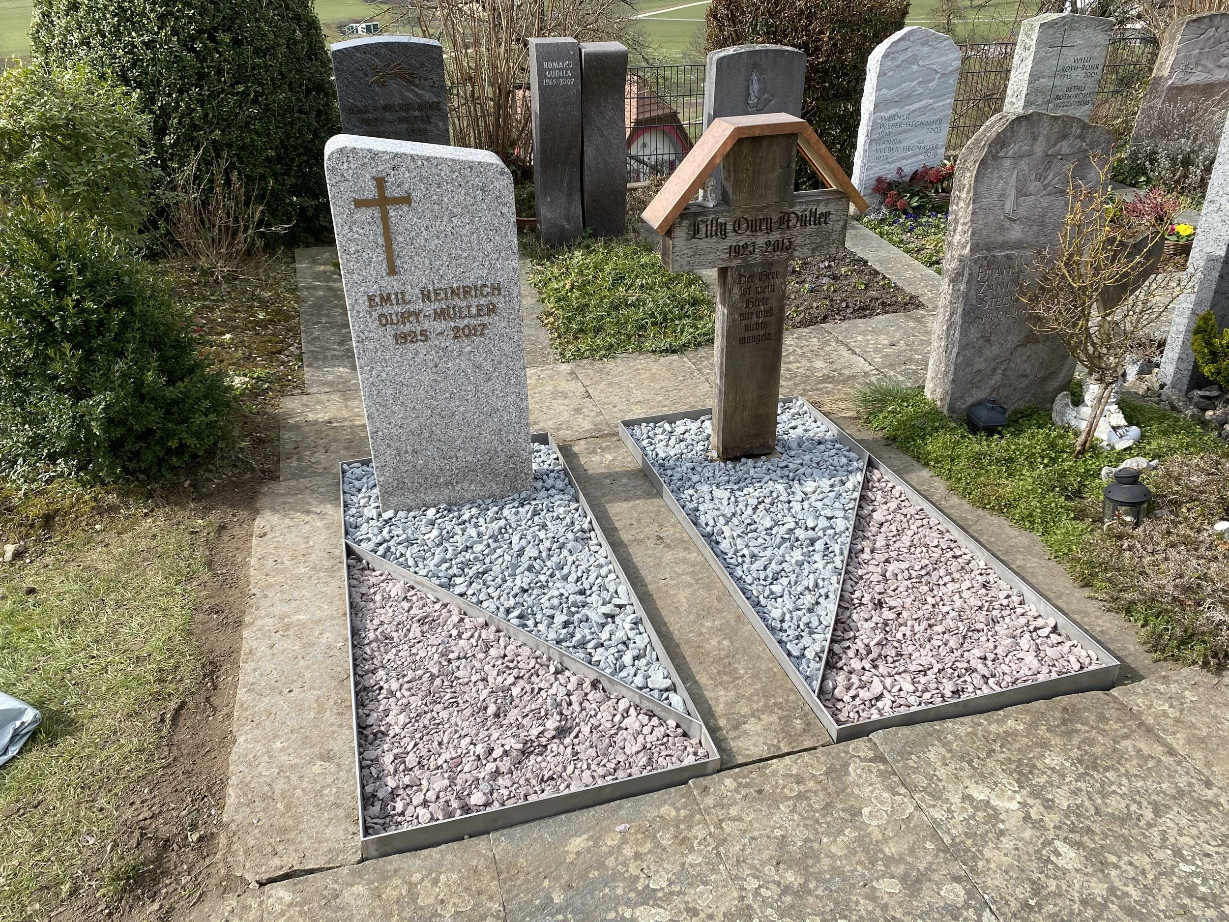 Zwei nahe beieinander liegende Grabsteine auf einem Friedhof, jeweils mit Dränagetrennbändern, der eine ist aus hellem Granit mit goldenem Kreuz, der andere aus dunklem Holz mit Dachüberstand. Es sind weitere Gräber und Pflanzen im Hintergrund sichtb