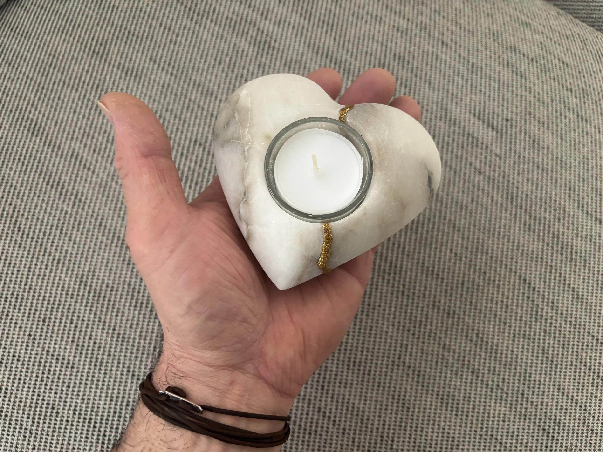 Hand hält eine Steinskulptur in Herzform mit einer Kerze in der Mitte, auf einem grauen Stoffhintergrund.