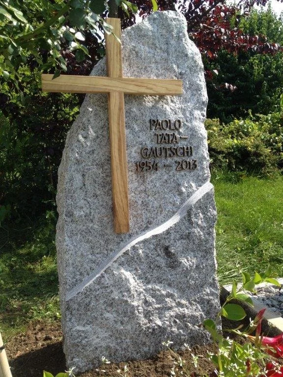Grabstein aus Stein mit Holzkreuz und eingravierter Inschrift, umgeben von Pflanzen.