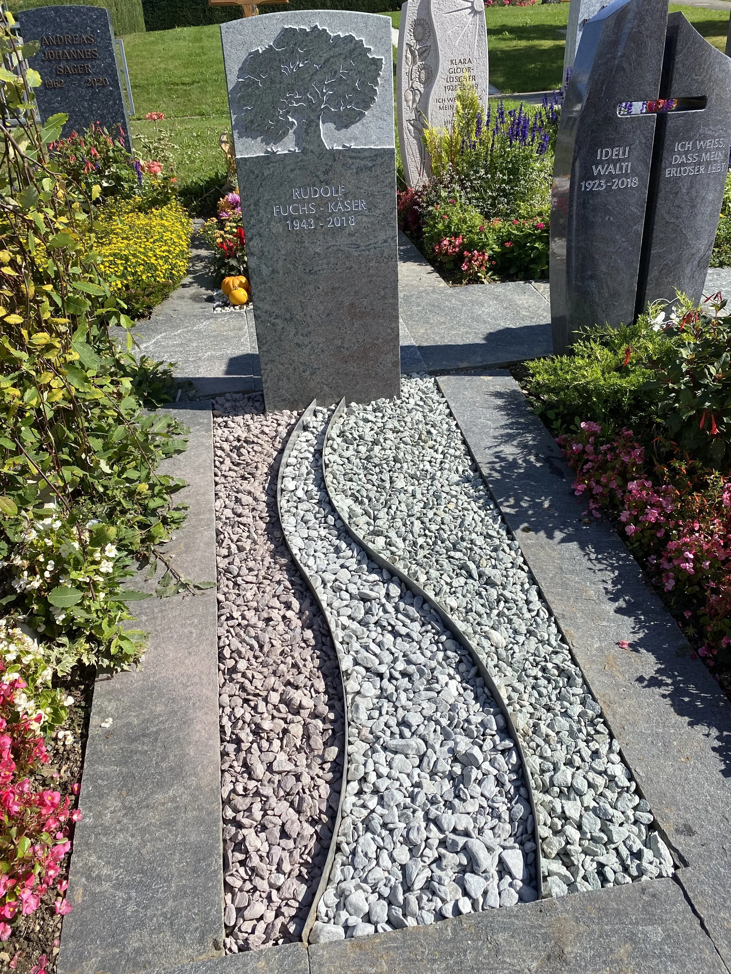 Ein Grab im Friedhof mit bepflanzten Blumen und Steinen, zwei Grabsteine mit Inschrift, einer mit einem Baum-Logo, der andere mit Sonnenblumen-Relief.