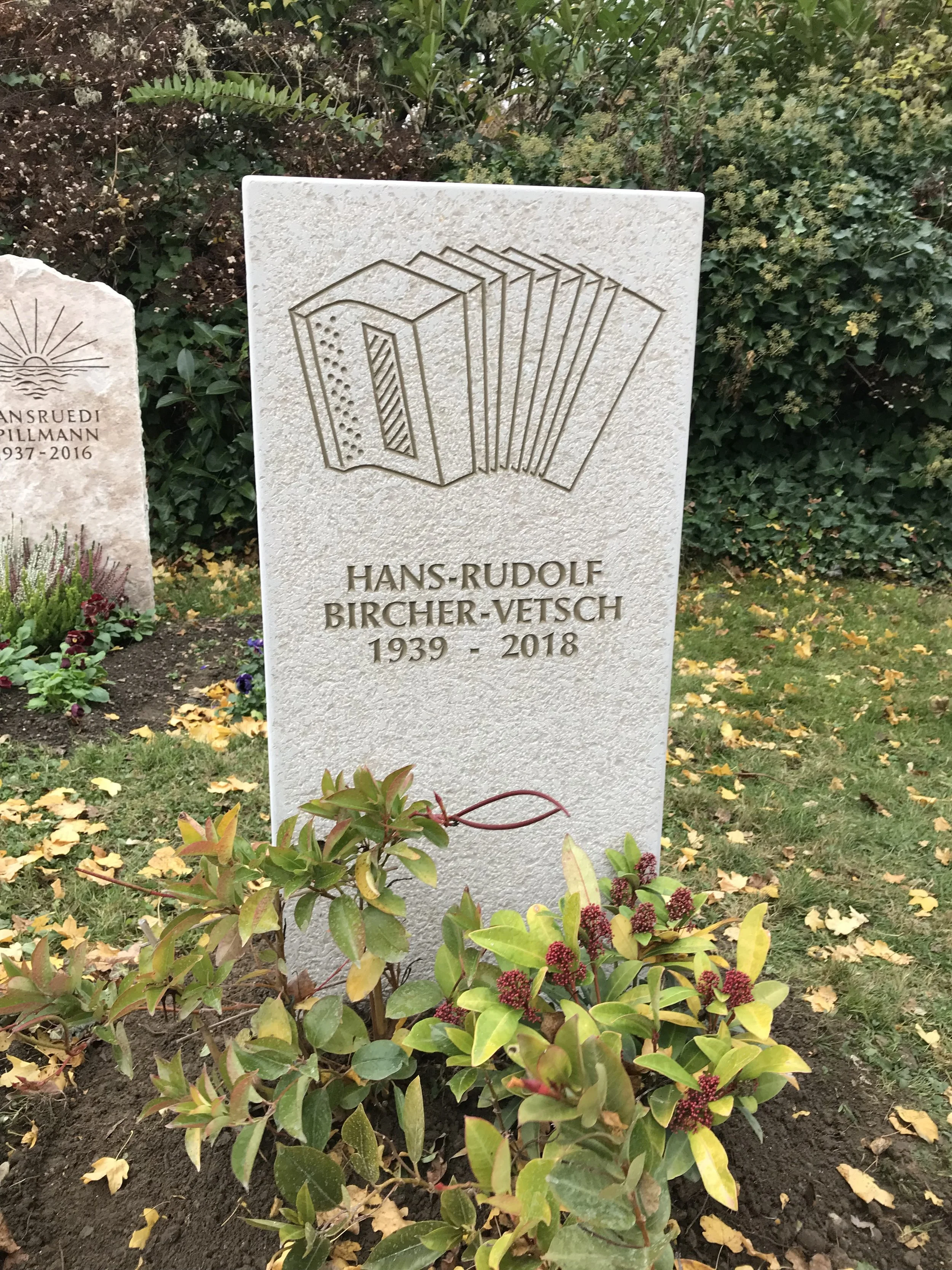 Grabstein mit einem Buch-Logo und dem Namen Hans-Rudolf Bircher-Vetsch, der 1939 bis 2018 lebte, in einem Garten mit anderen Gräbern und blühenden Pflanzen.