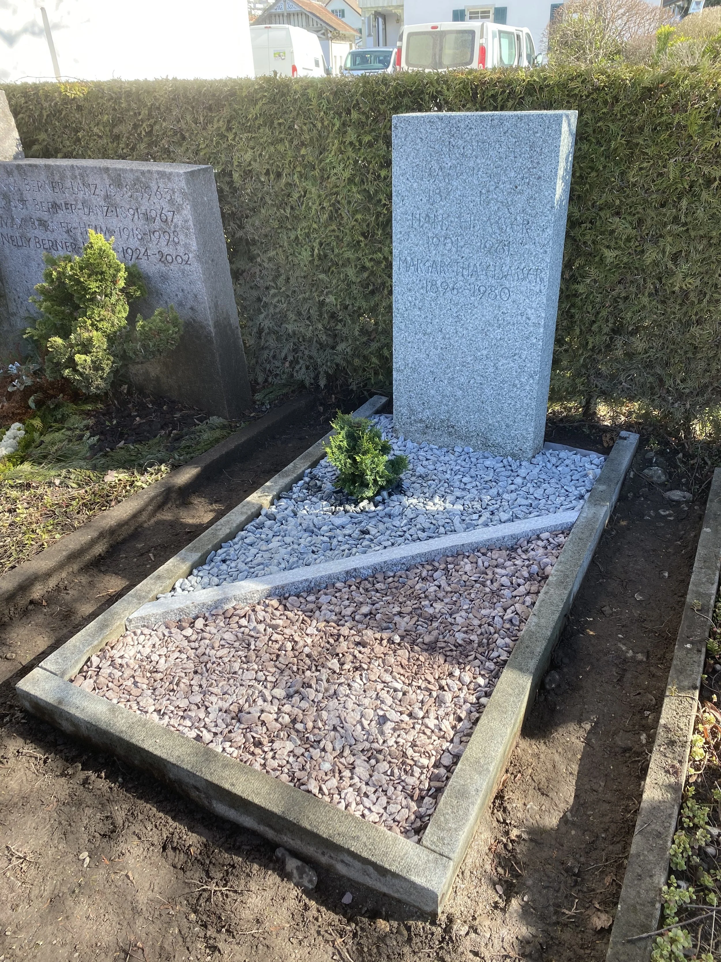 ein frisch gepflegtes Grab mit zwei Grabsteinen, einem kleinen Strauch und einem Bereich mit Kieselsteinen, umgeben von Büschen und Zäunen.