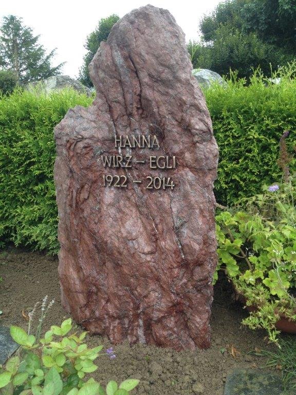 Stein mit gravierter Inschrift, umgeben von Pflanzen, im Hintergrund Bäume und ein bewölkter Himmel.