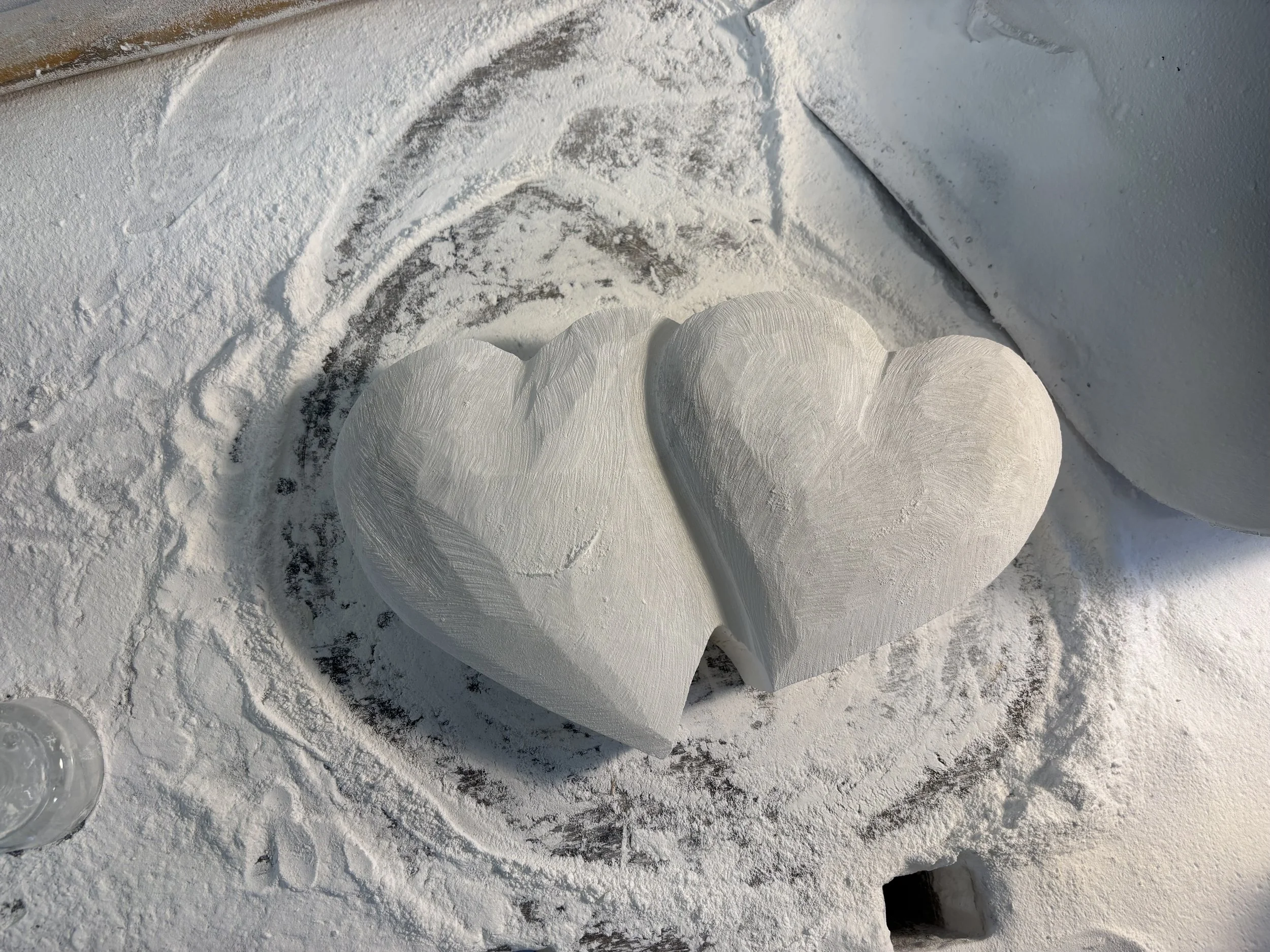 Zwei weiße, herzförmige Skulpturen, die aus Holz geschnitzt sind, liegen auf einer schneeüberdeckten Oberfläche, umgeben von Schnee und einer langen Linie aus Staub oder Puder.