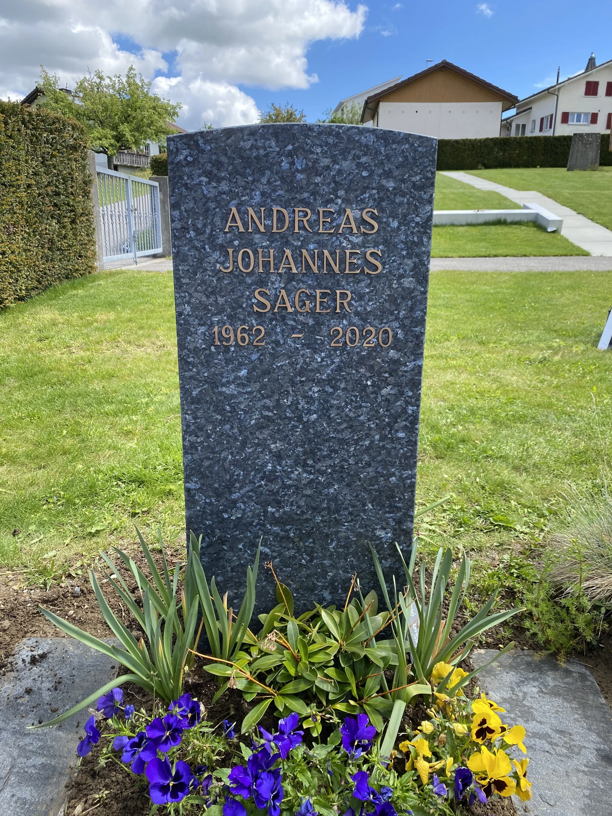 Grabstein mit dem Namen Andreas Johannes Sager, 1962-2020, auf einem Friedhof, umgeben von Blumen und grünem Gras, mit Himmel und Himmel im Hintergrund.