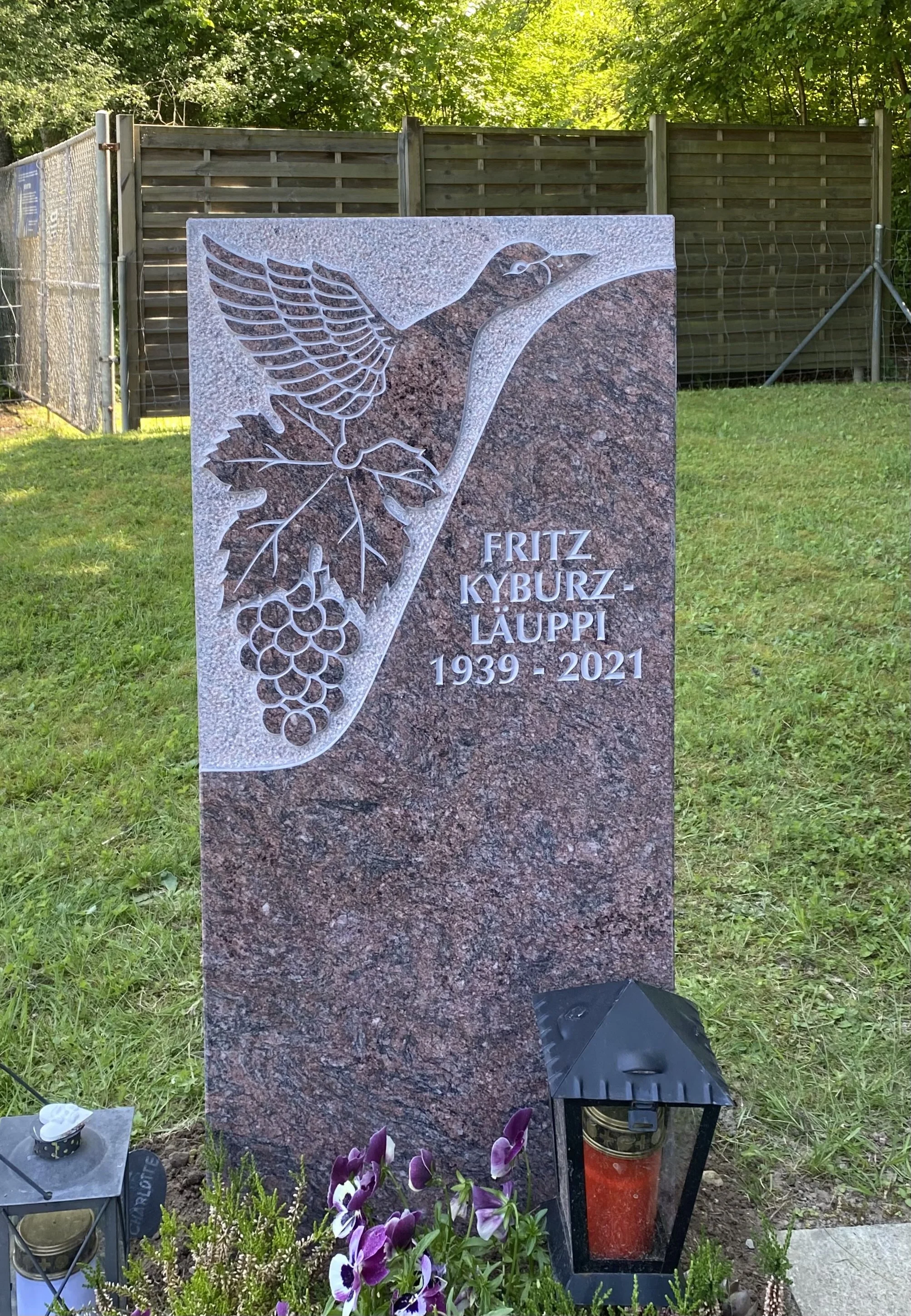 Grabstein mit eingraviertem Vogel, Trauben und Blättern, der Text lautet: Fritz Kyburz-Laupp 1939-2021, auf dem Grab liegt eine kleine Laterne und Blumen.
