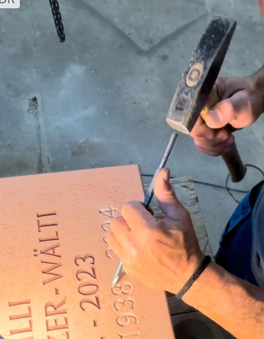 Ein Handwerker arbeitet mit einem Hammer und einem Meißel auf einem Stein, auf dem eingravierte Daten und Buchstaben sind.