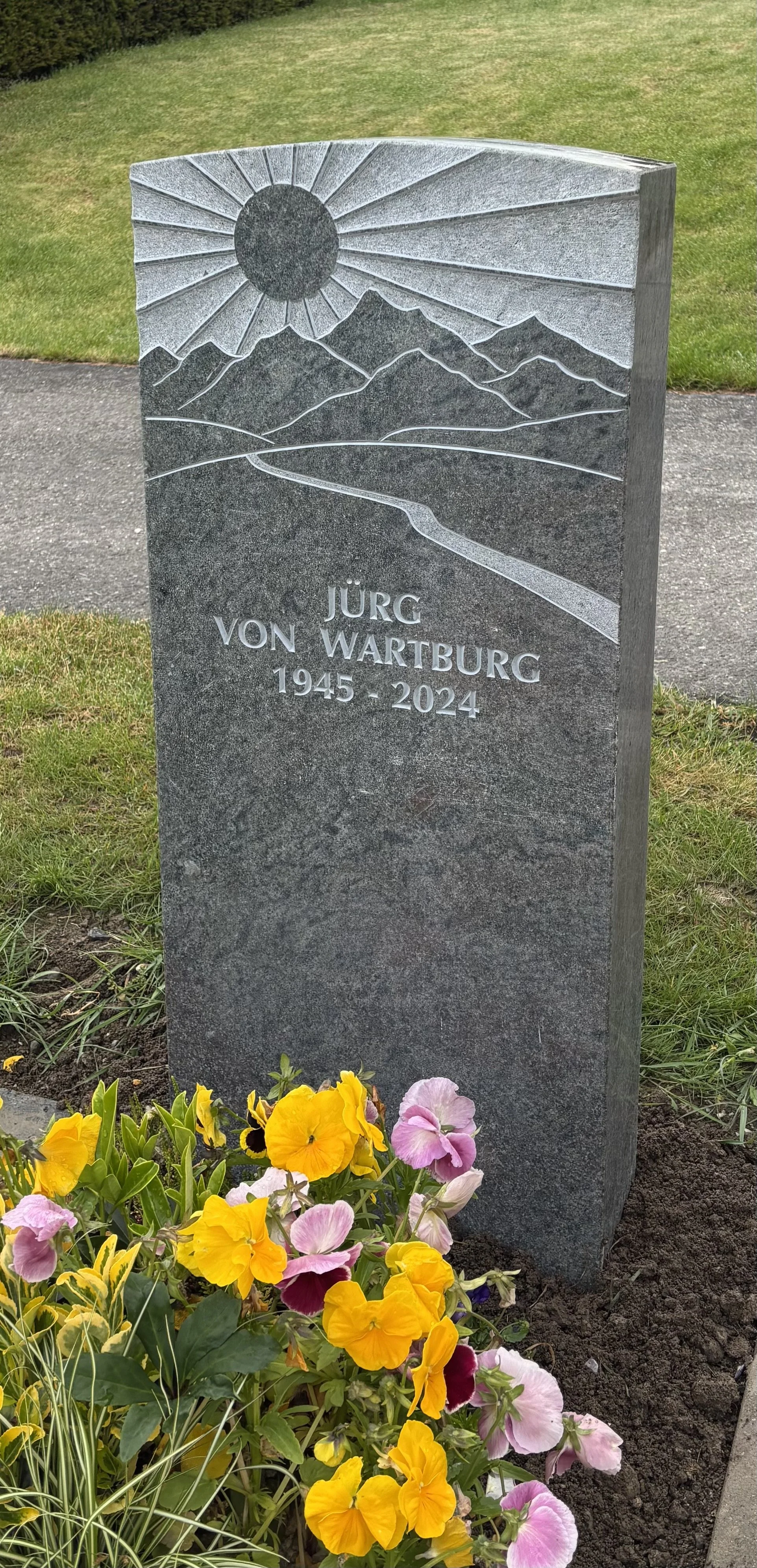 Grabstein mit Sonnen- und Bergmotiven, Name Jürg von Wartburg, Geburtsjahr 1945, Sterbejahr 2024, Blumen vor dem Grab.