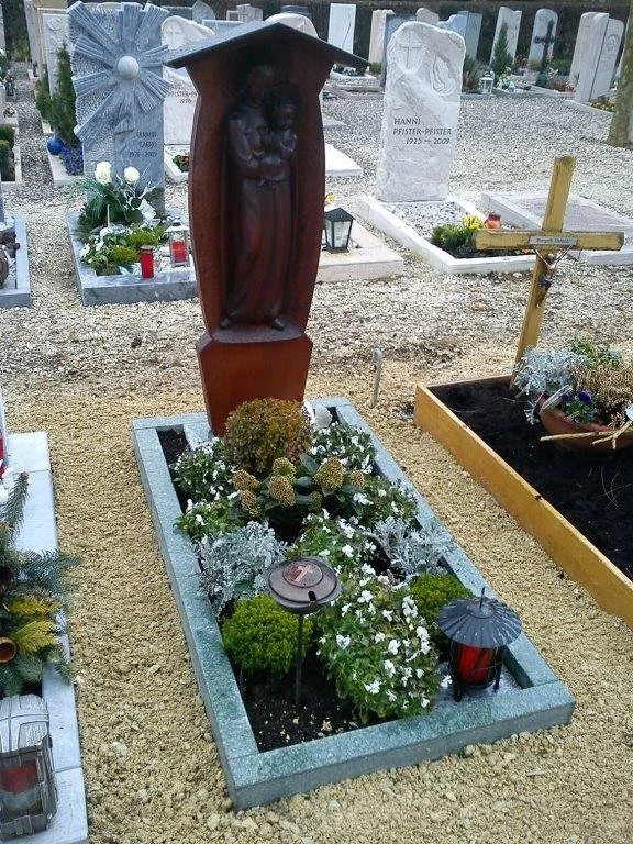 Grabhügel mit Holzkreuz und Holzfigur einer Mutter mit Kind, Blumen, Kerzen und Dekorationen auf einem Friedhof.