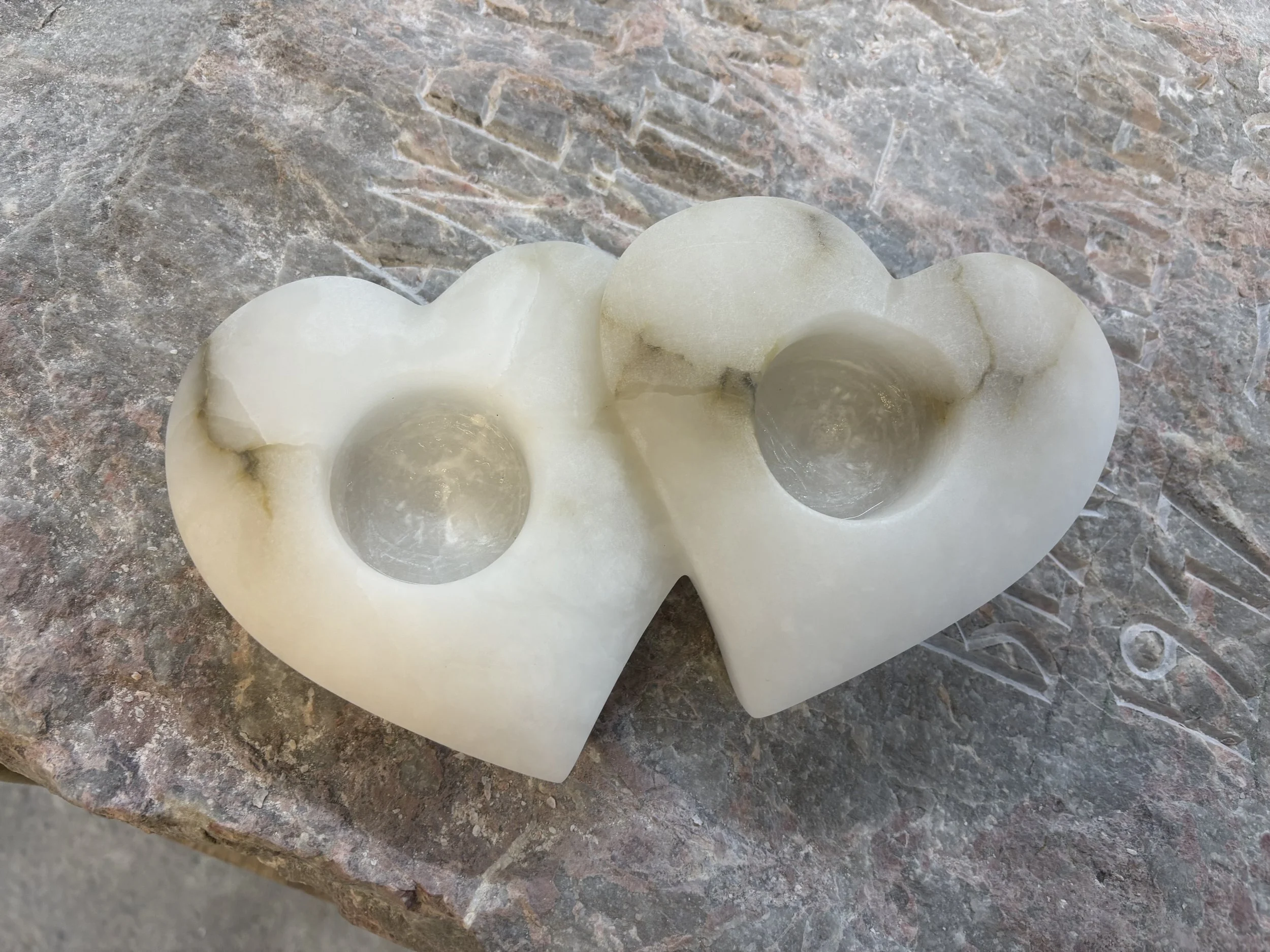 Eine weiße, herzförmige Kerzenhalter aus Marmor mit zwei runden Kerzenhaltern, liegt auf einem grauen, strukturierten Steinuntergrund.