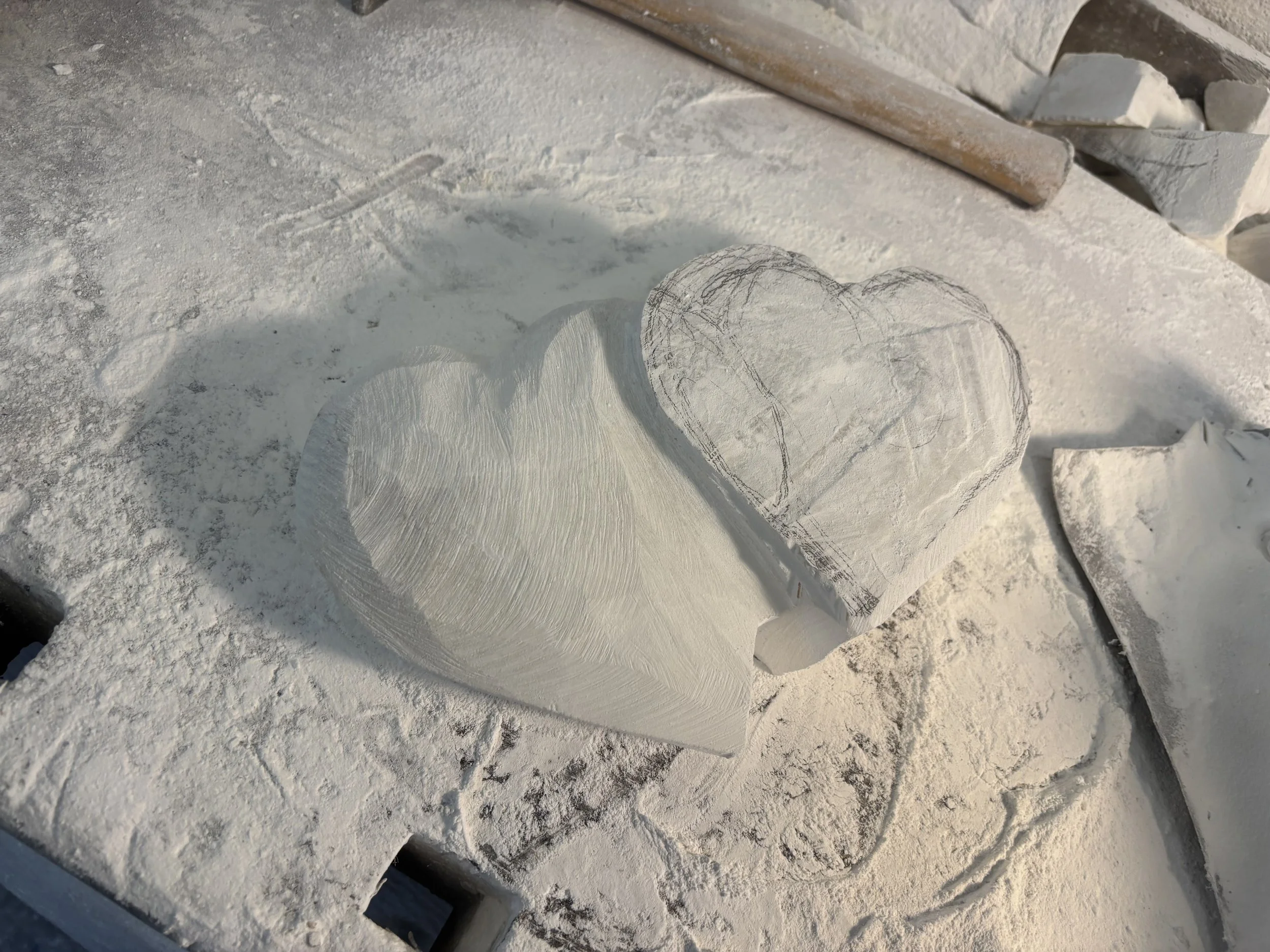 Zwei Herzen aus Holz, eines fertig gefertigt und eines noch im Umriss, liegen auf einem Schneideblock, umgeben von Holzspänen und Werkzeugen.