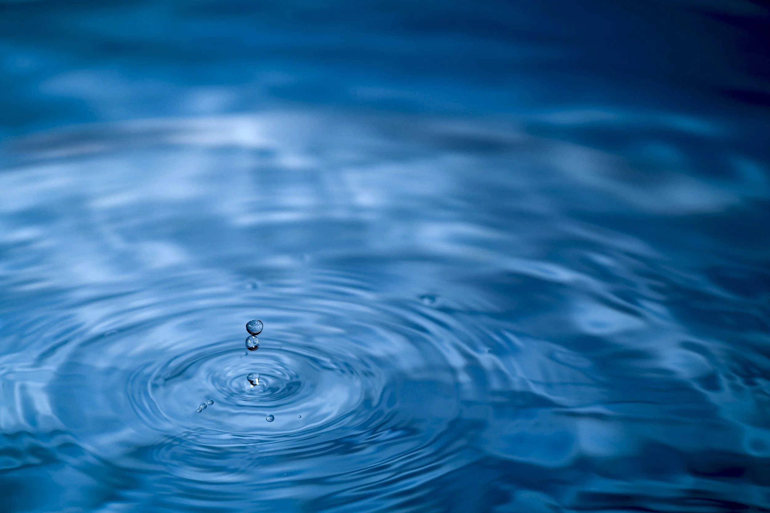 Close-up of a water droplet falling into a calm blue body of water, creating ripples and bubbles.