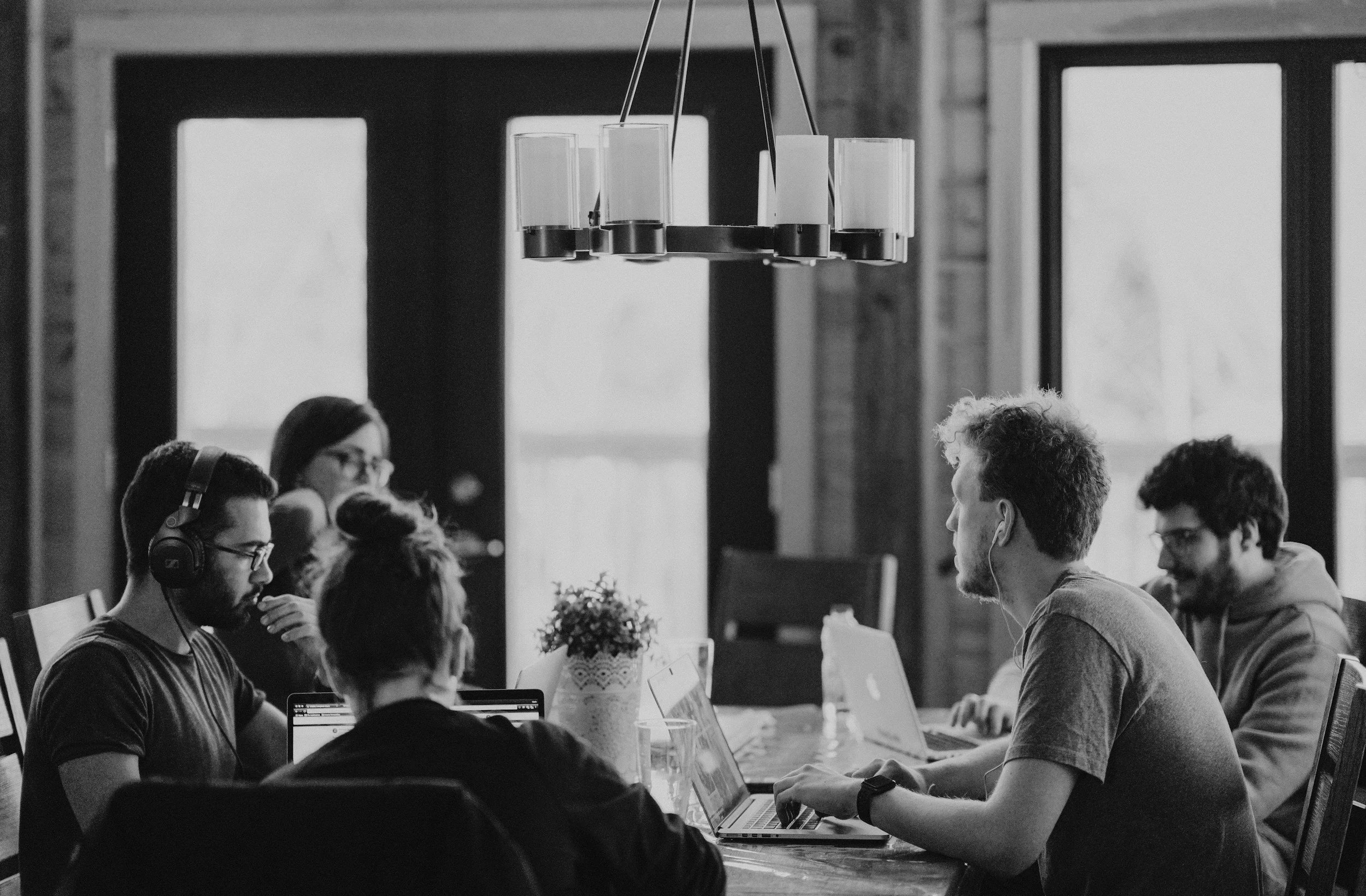 A group of five people working on laptops around a wooden table in a modern office with large windows, a hanging light fixture, and a potted plant in the center.