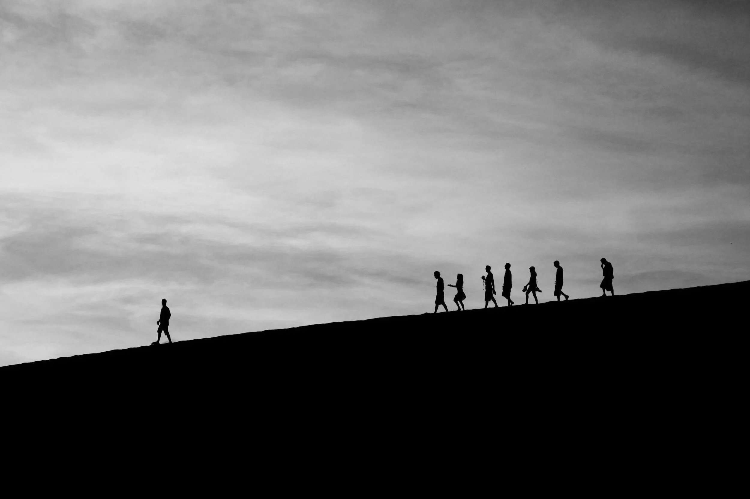 Silhouette of a person leading a group walking on a hilltop during sunset or sunrise, with a cloudy sky in the background.