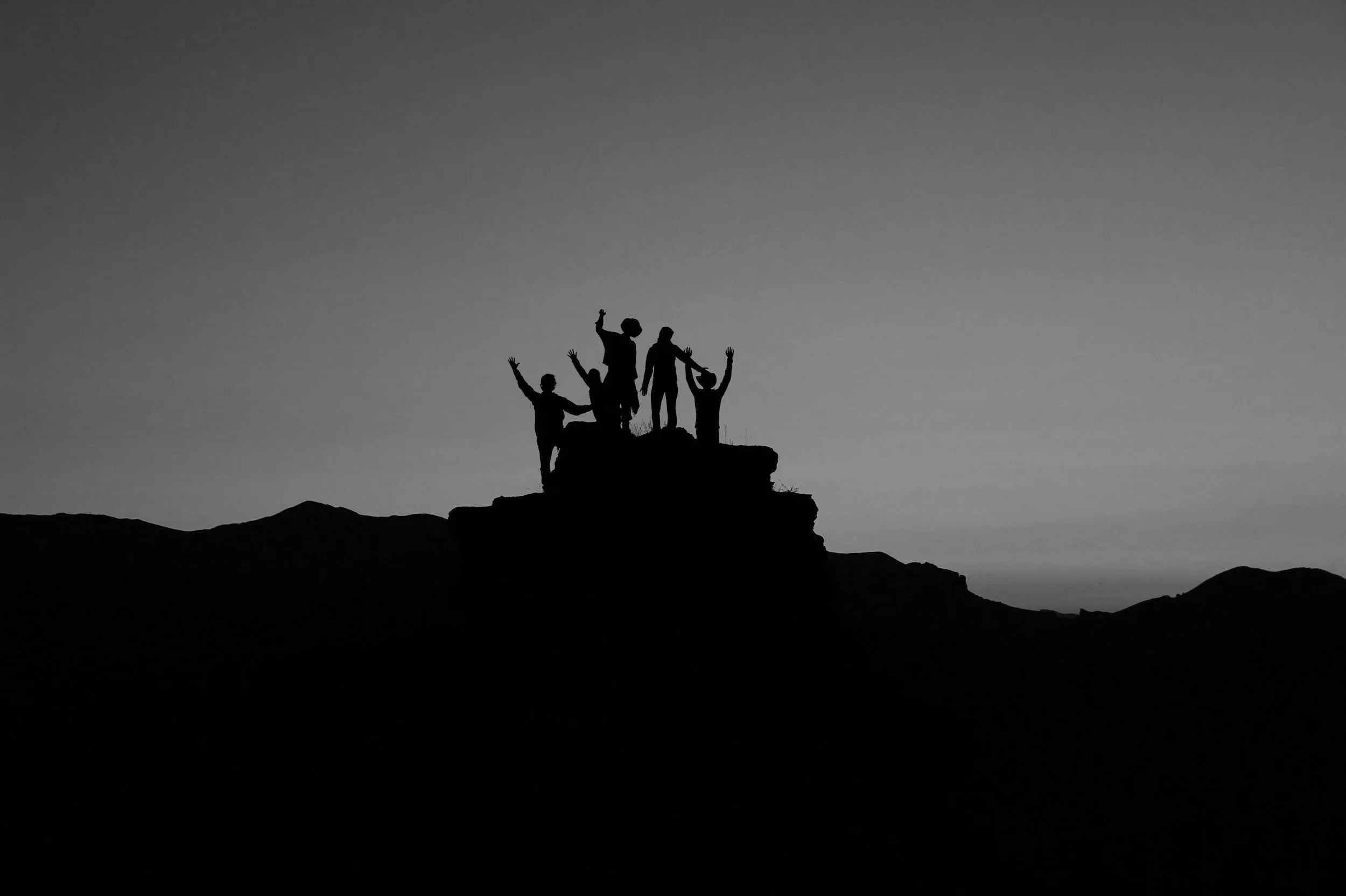 Silhouettes of five people on a rocky hilltop, with some raising their arms, against a dusky sky.