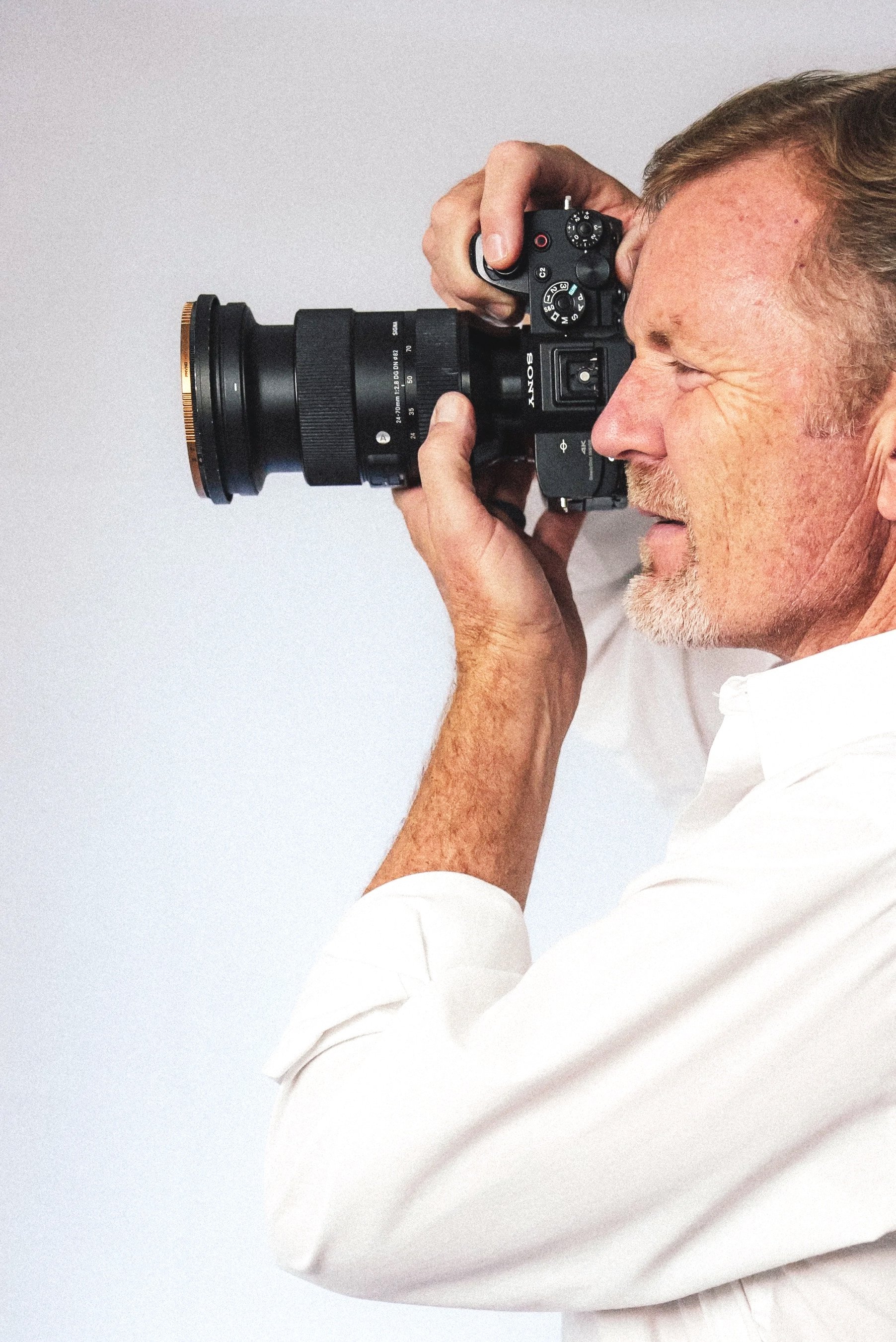 A man with gray hair and a beard wearing a white shirt, taking a photograph with a black Sony camera, against a plain light background.