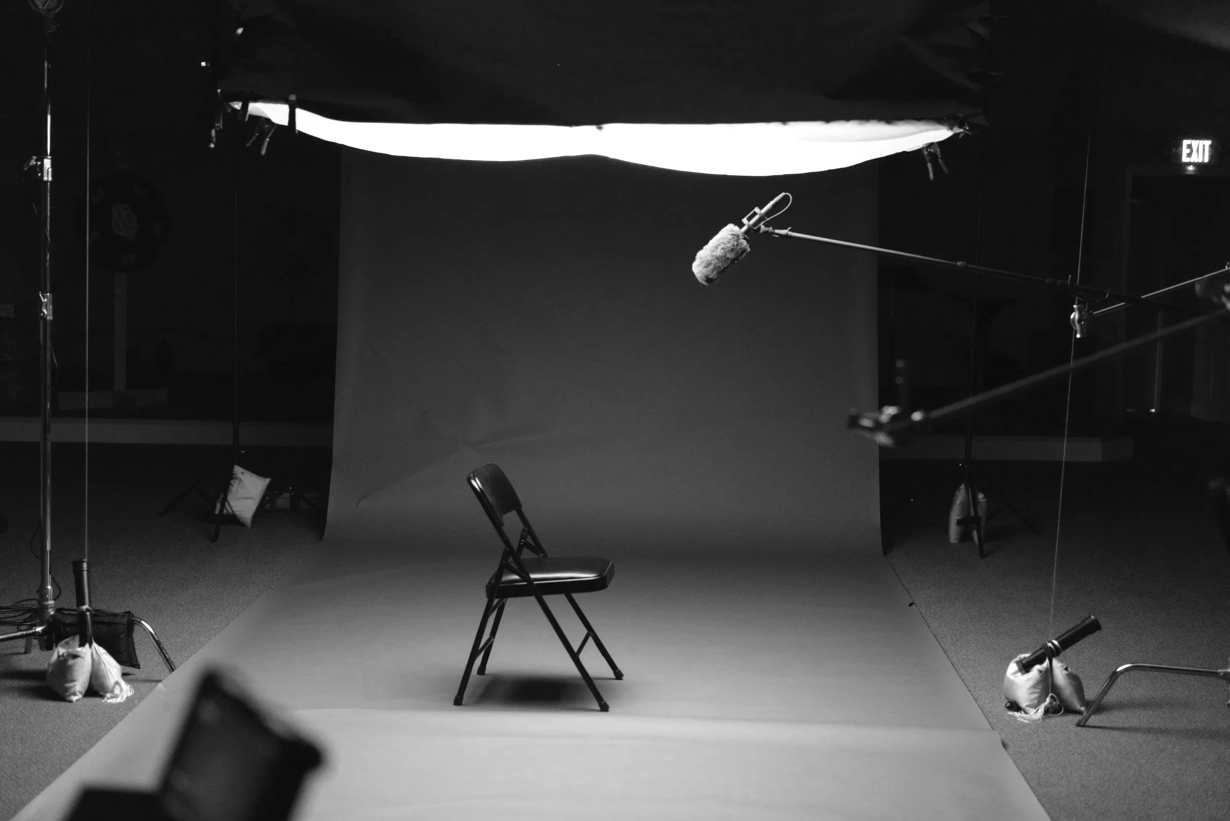 Empty chair in front of a gray seamless background, surrounded by lighting equipment, a boom microphone hanging overhead, and other film production accessories in a studio setup.