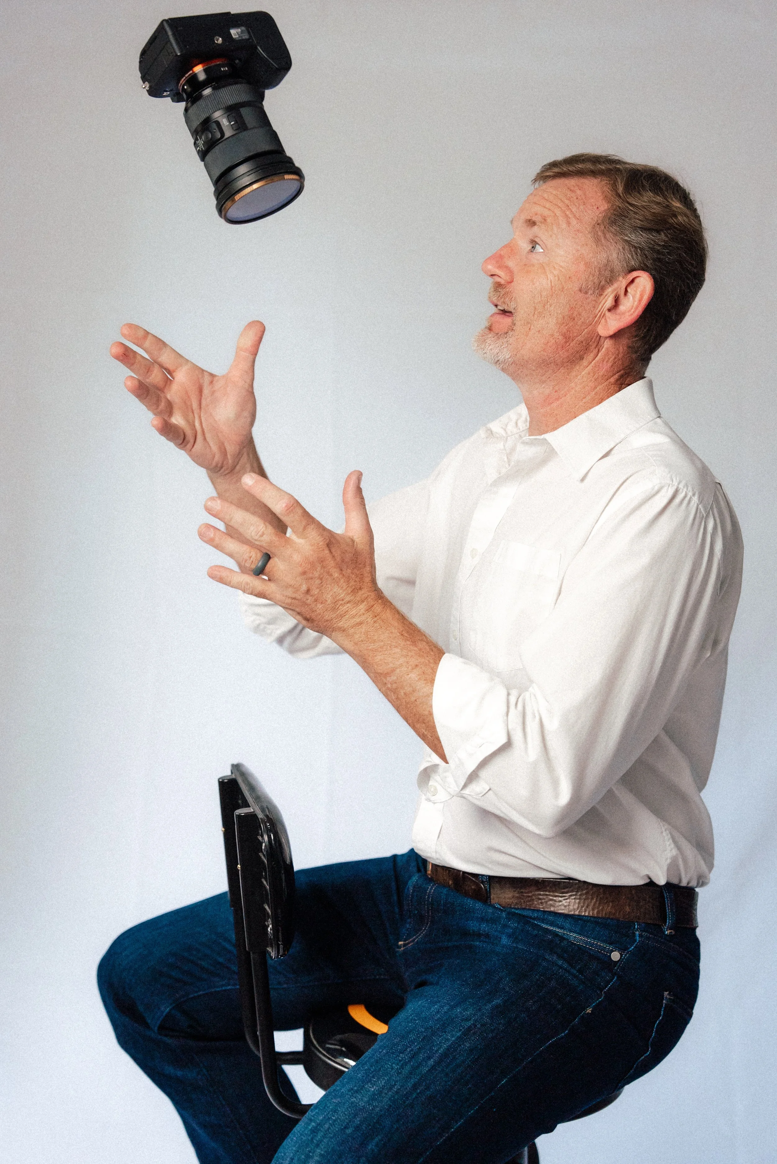 A man in a white shirt and blue jeans is sitting on a stool, tossing a camera in the air and watching it with excitement.