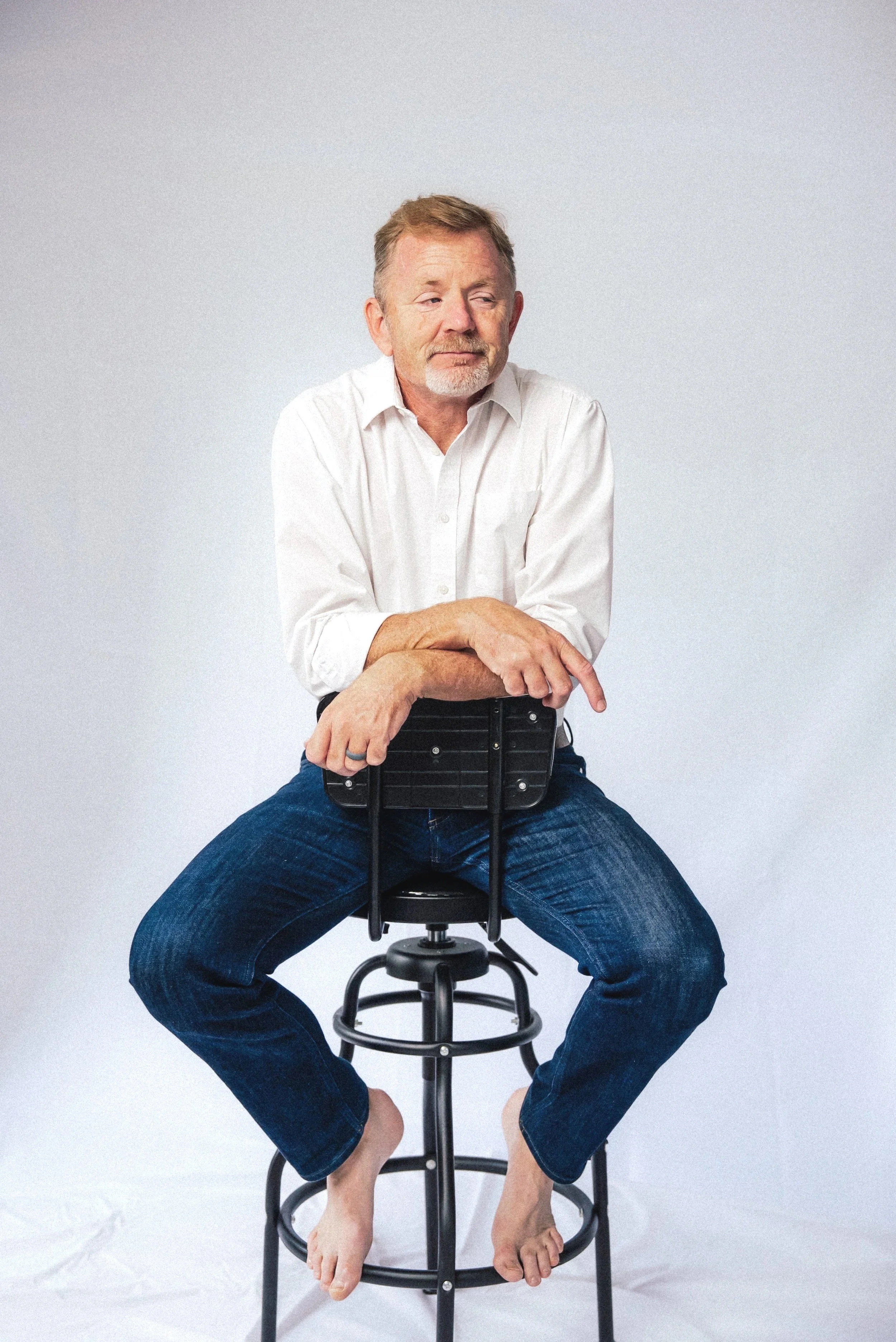 A middle-aged man with gray hair and beard sitting barefoot on a black stool, wearing a white shirt and jeans, against a plain white background.