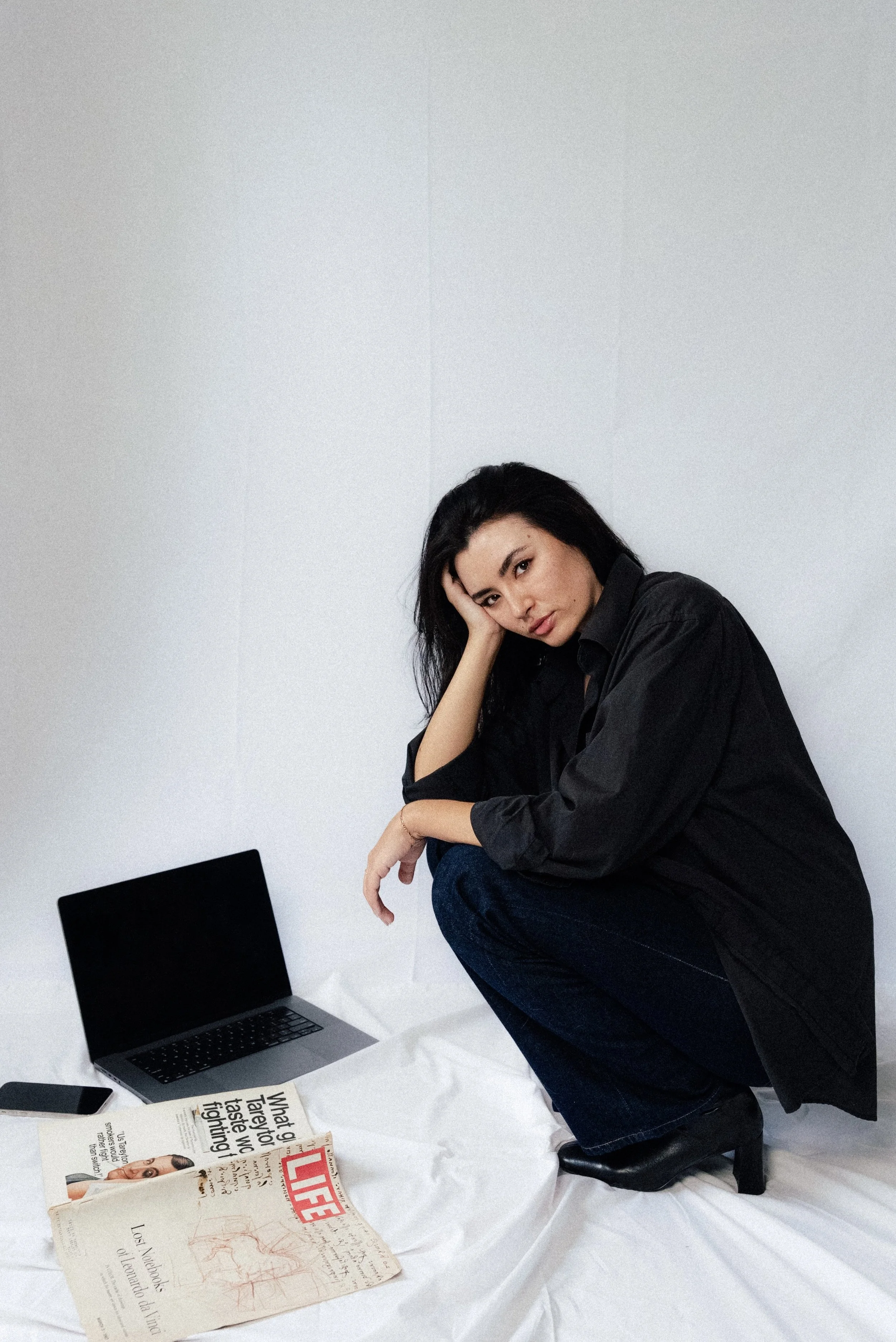 A woman with black hair in a black shirt and jeans squats on a white surface with a white wall background. She rests her head on her left hand and looks at the camera. There is a laptop, a smartphone, and newspapers on the surface.