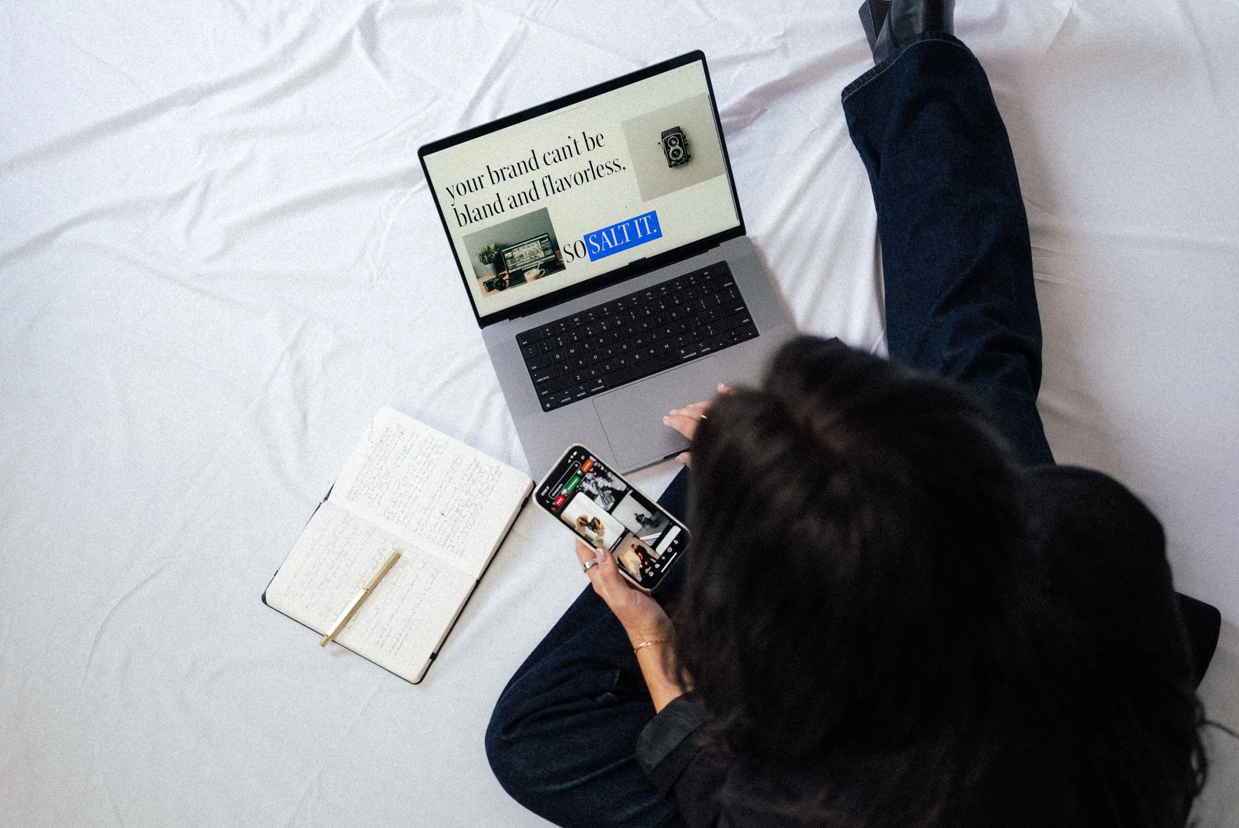 A person sitting on a bed using a laptop and phone, with a notebook nearby, on a white bedsheet.
