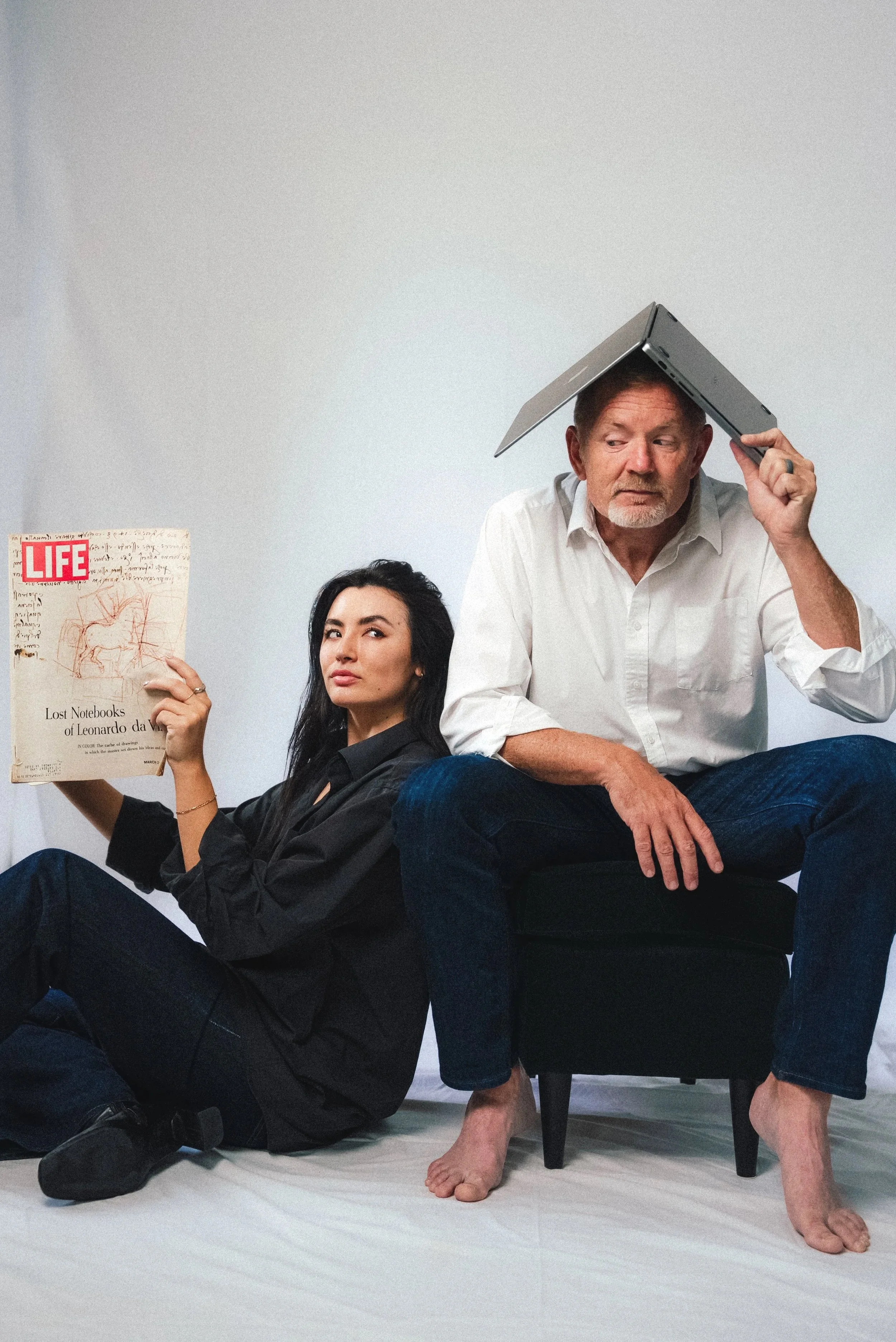 A young woman sitting on the floor leaning against a wall, holding a vintage LIFE magazine featuring Leonardo da Vinci's sketches. An older man sitting on a small black chair with a laptop on his head, appearing to balance it, wearing a white shirt and jeans, with one bare foot resting on the ground.