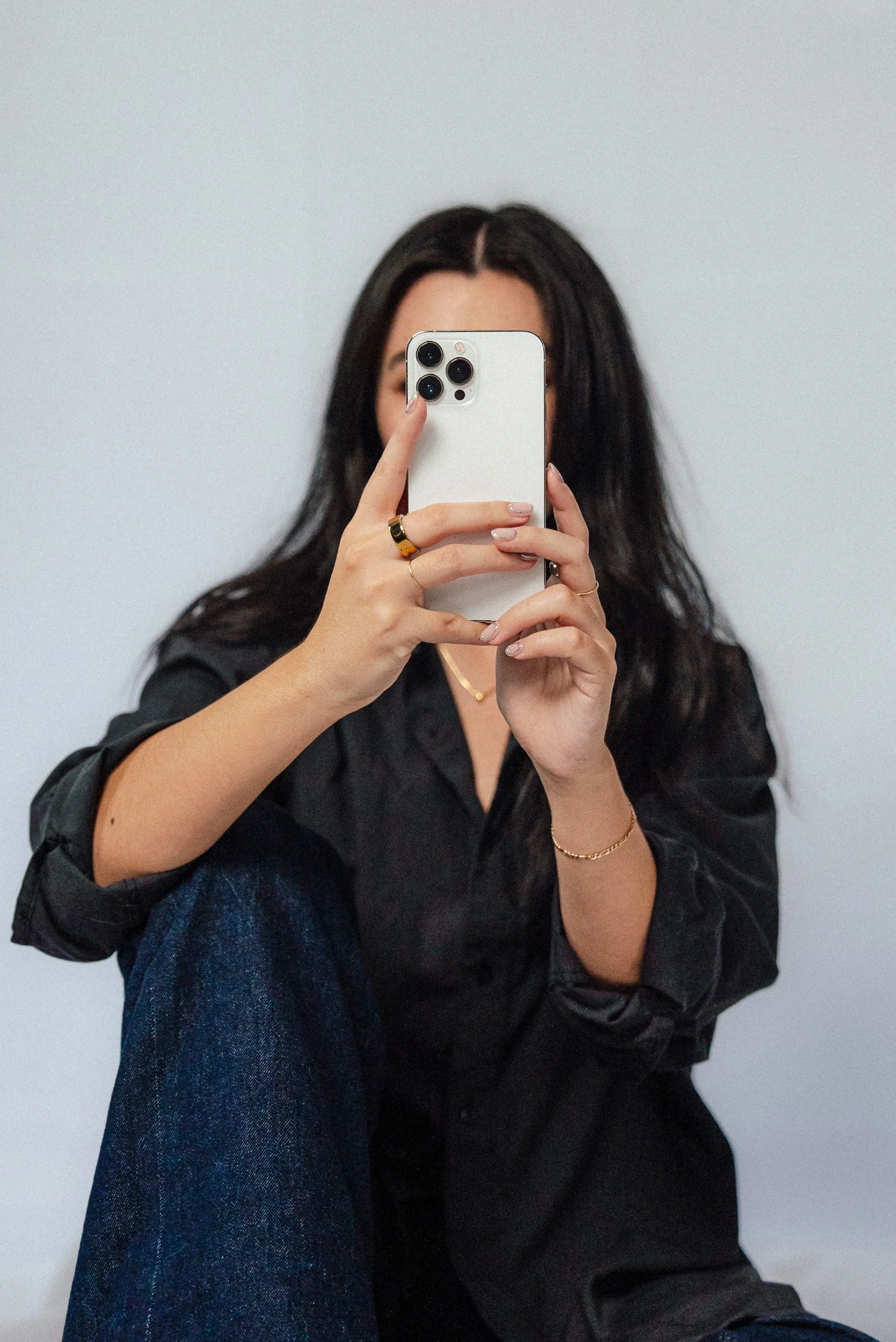 A person with long dark hair taking a mirror selfie with a white smartphone, sitting against a plain light gray wall, wearing a black shirt, blue jeans, and gold jewelry posting to social media