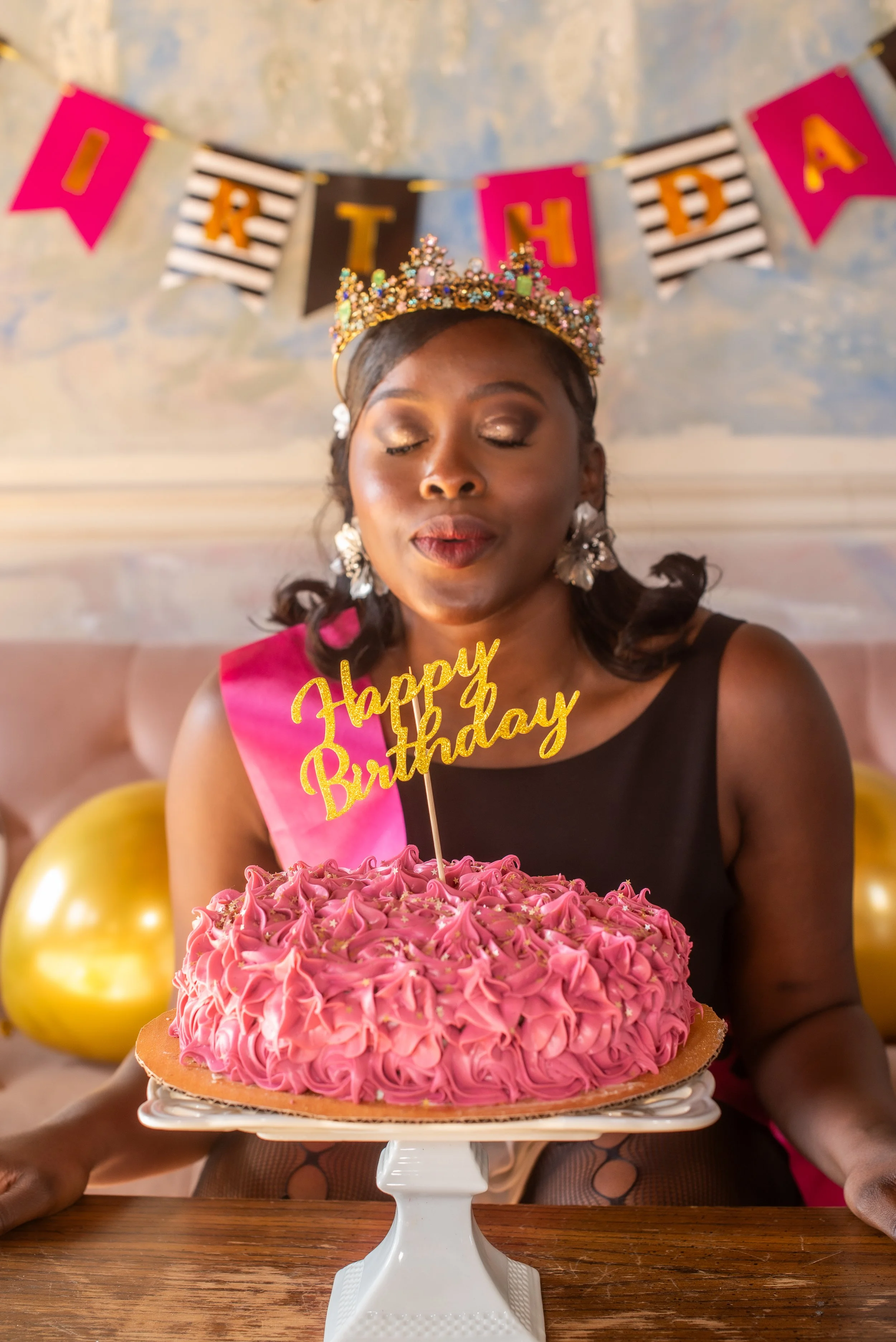 Black woman blowing out candles on a pink and gold birthday cake