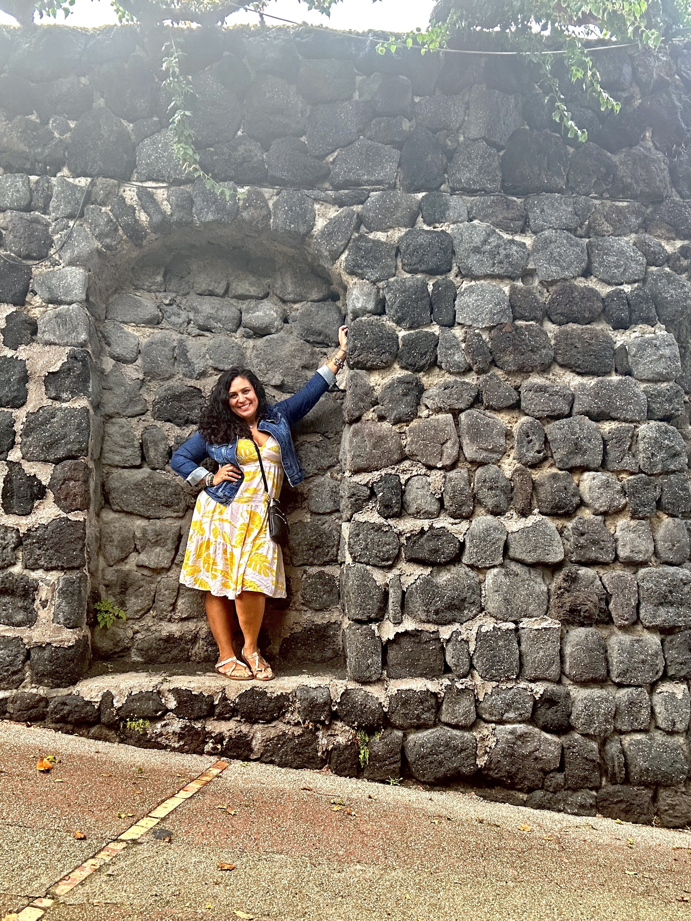 Italian woman standing in front of stone wall in yellow dress and sandals