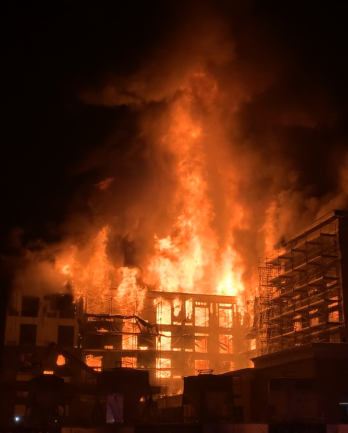 A large building fire with intense flames and thick smoke at night, with scaffolding visible on the side of the structure.