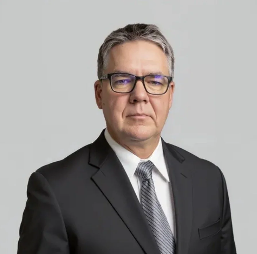 A middle-aged man with gray hair and glasses wearing a black suit, white shirt, and striped tie, standing against a plain light gray background.