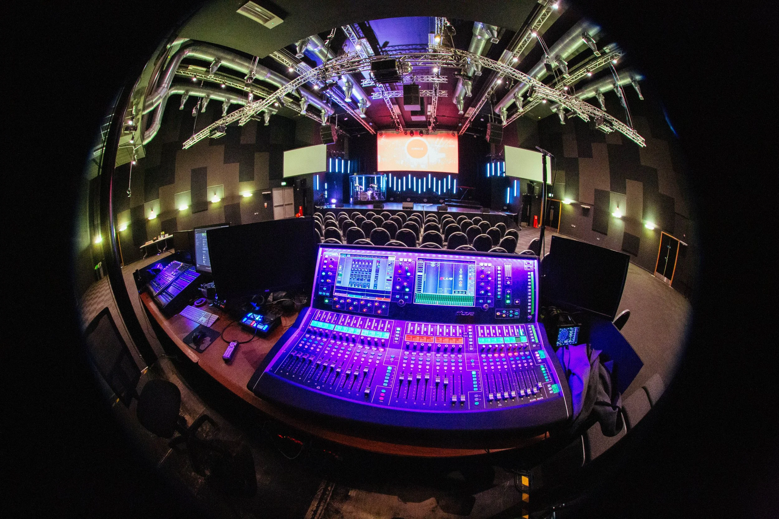 Empty theater or concert venue with a stage and chairs, viewed from the control booth which has a large sound mixing board with multiple screens and controls, colorful lighting.