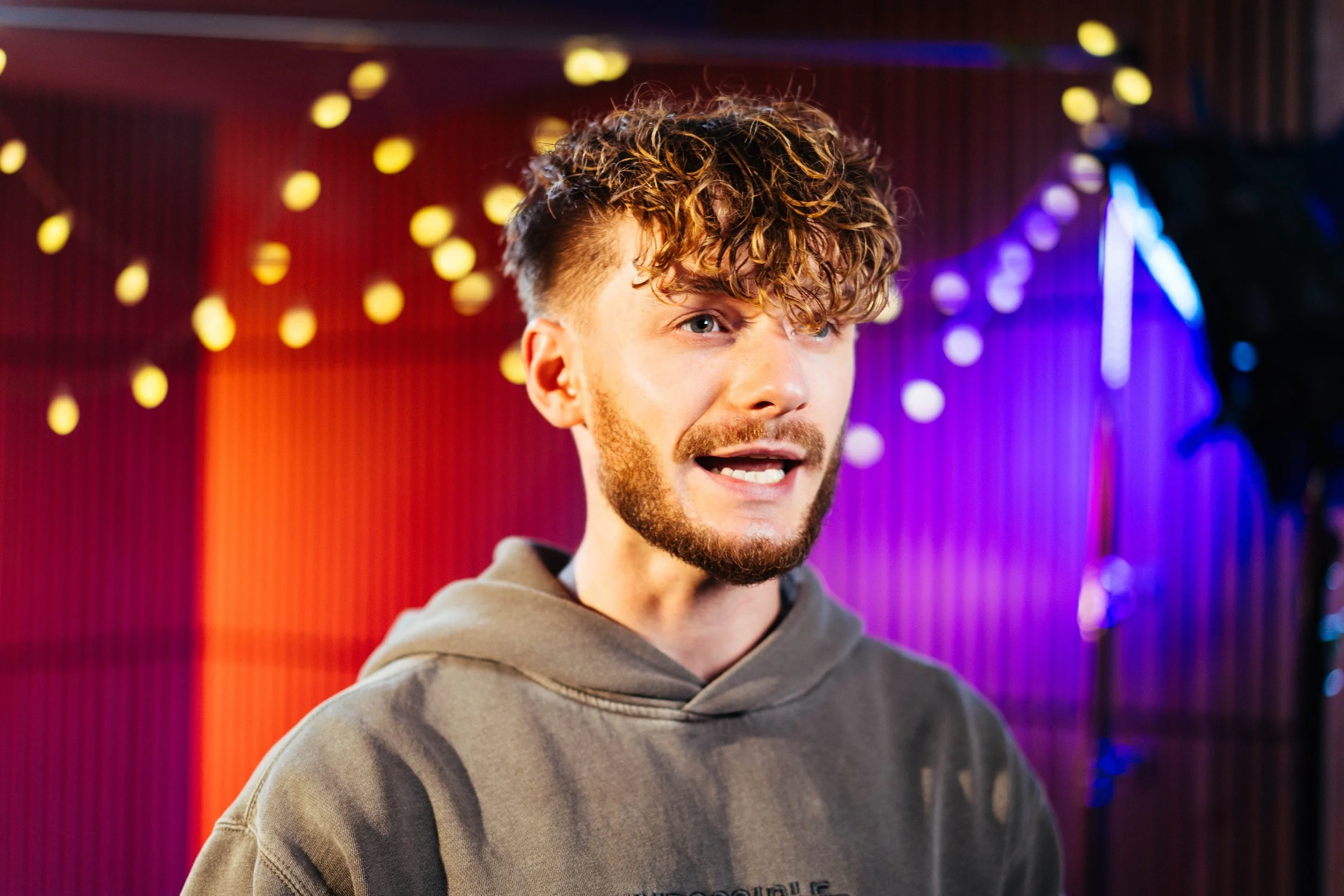 A young man with curly hair and a beard speaking or singing in front of a colorful background with string lights.