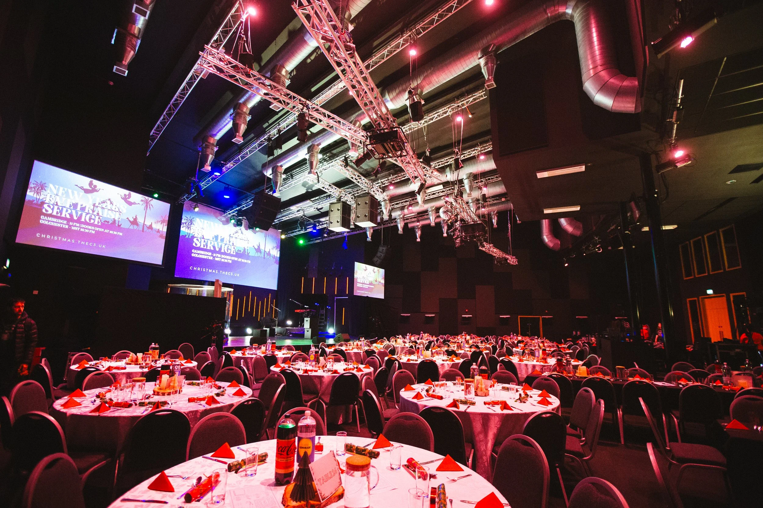 A large event hall decorated with round tables set for a Christmas celebration, featuring Christmas crackers, bottled drinks, and red napkins. The stage at the front displays large screens with a festive graphic and text for a Christmas service. The 