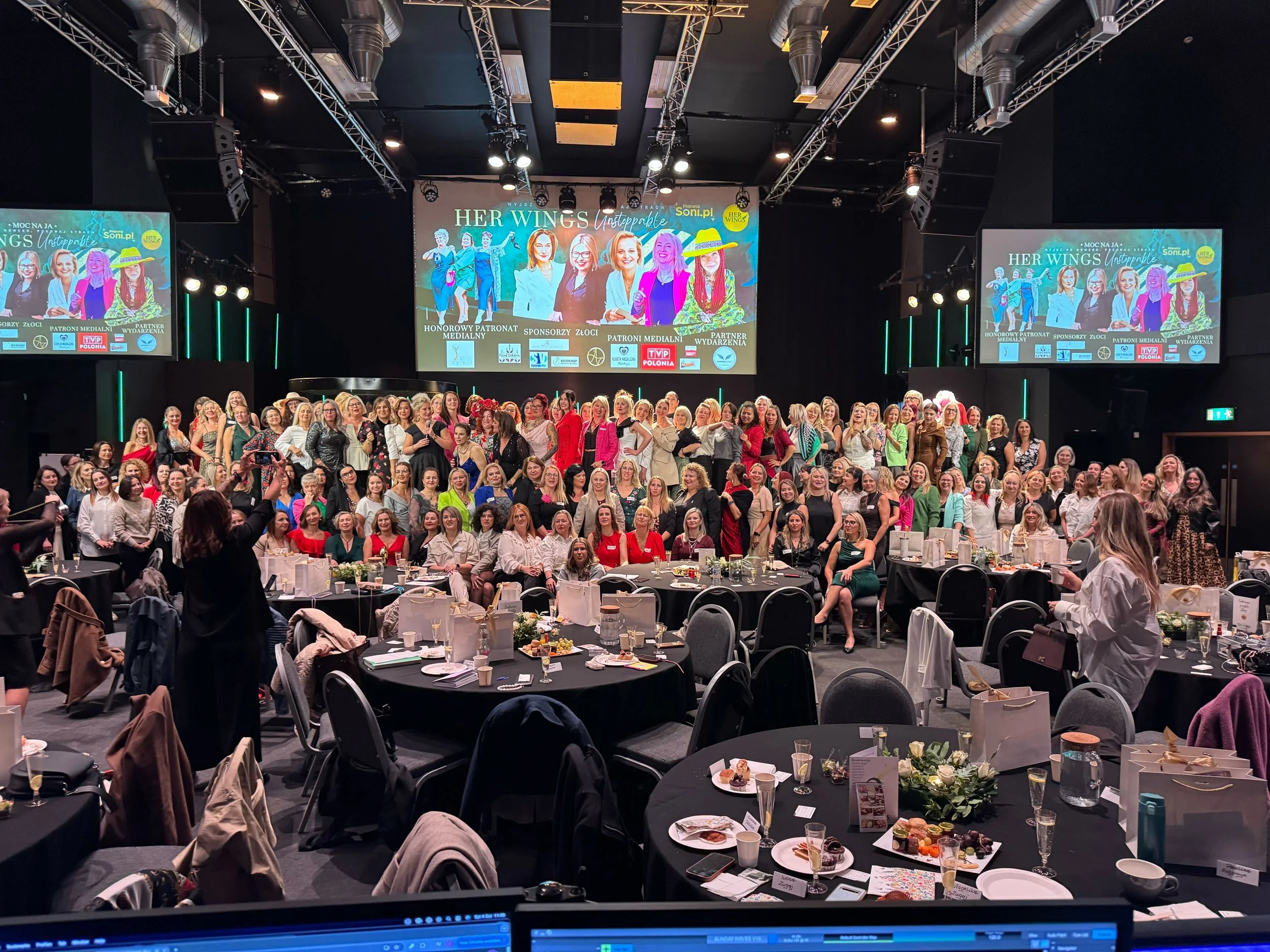 Large group of women gathered on stage and seated around tables at an event, with a large screen behind displaying the event's banner 'HER WINGS Unstoppable'. The tables have plates, glasses, and decorations, indicating a celebration or conference.