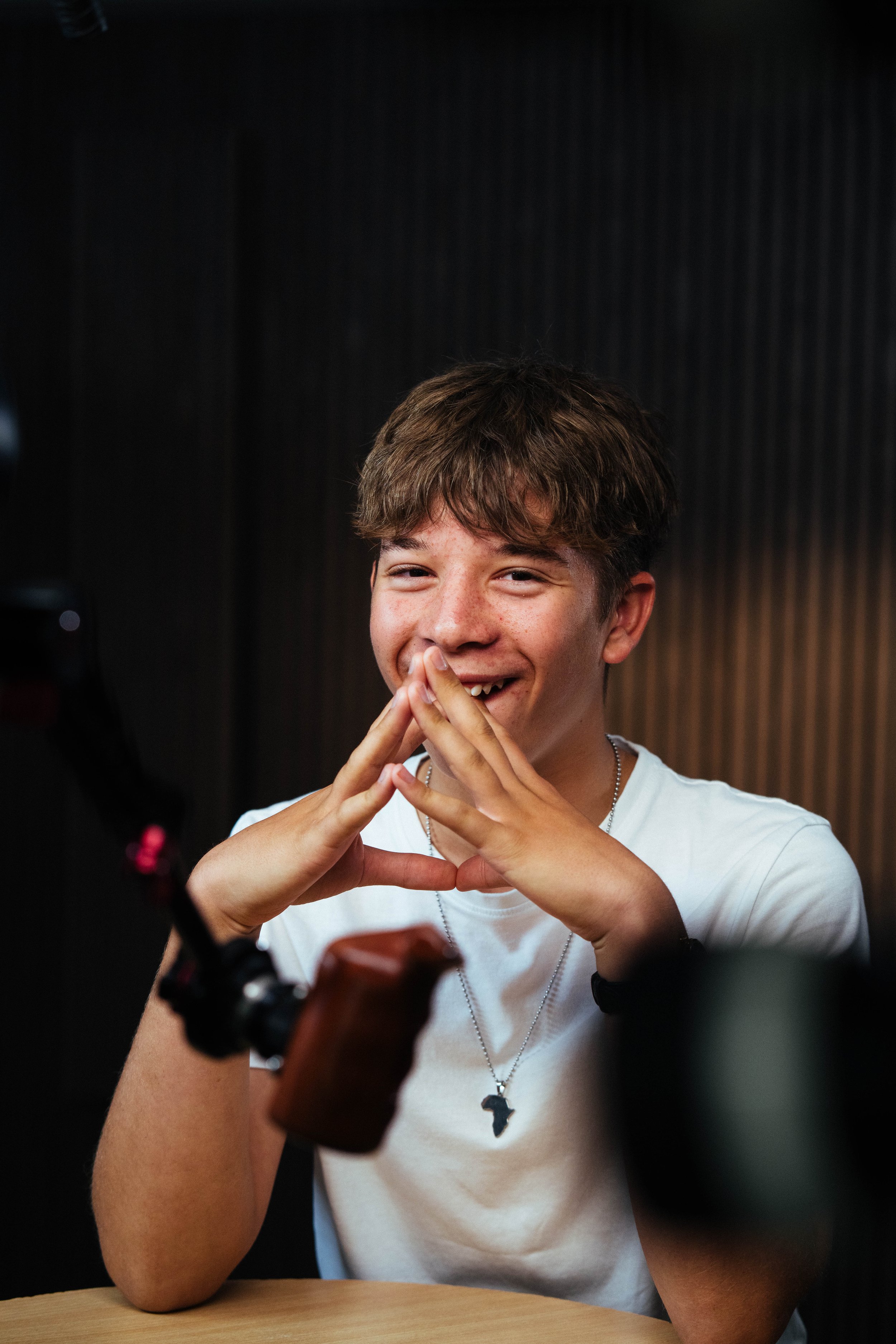 Smiling young man with short brown hair, wearing a white T-shirt and a necklace with a dark Africa-shaped pendant, sitting at a table in front of a camera.