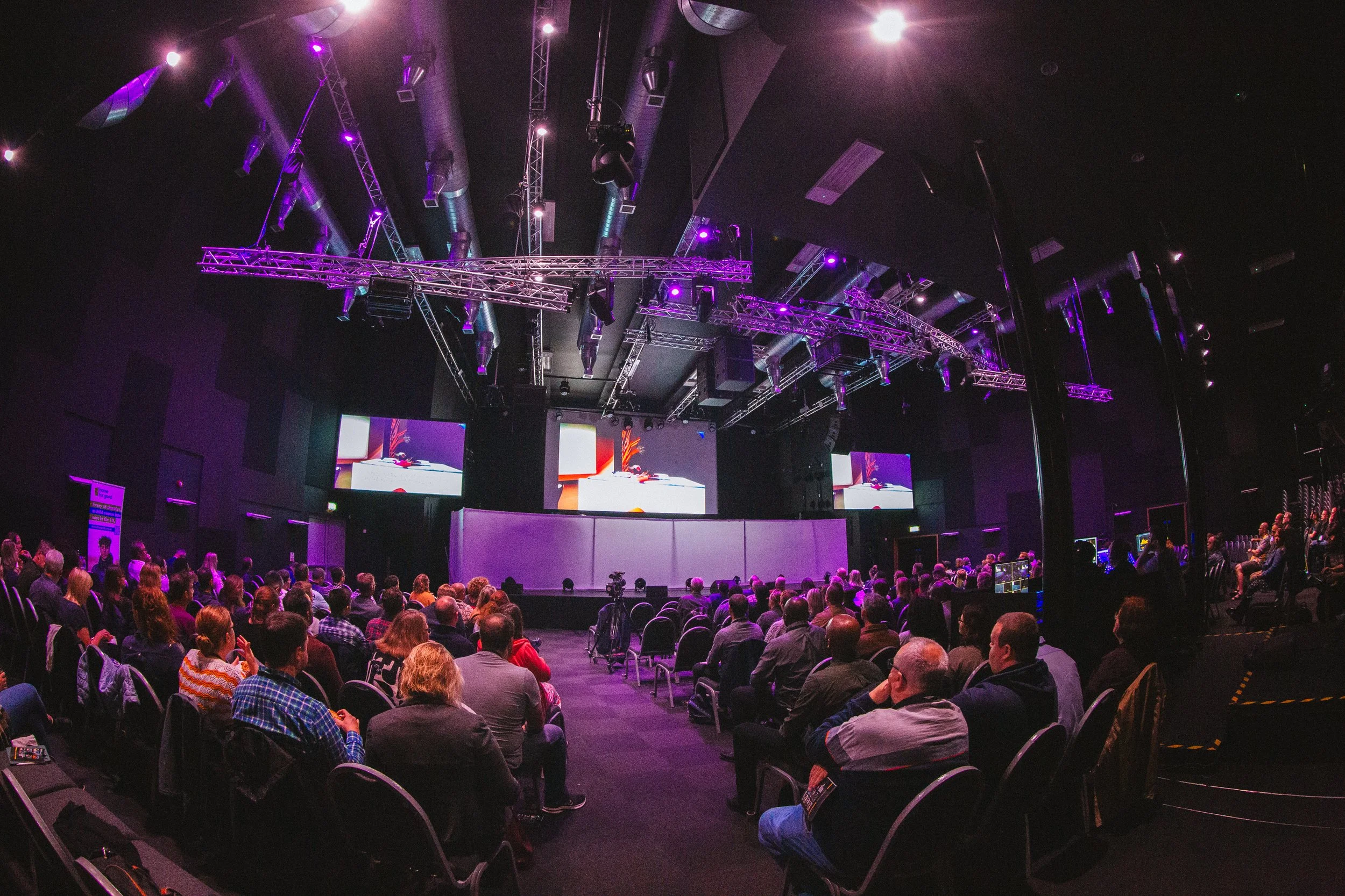 An indoor auditorium with a stage, large screens, and an audience seated watching a presentation or event. Purple and blue stage lighting illuminates the scene.
