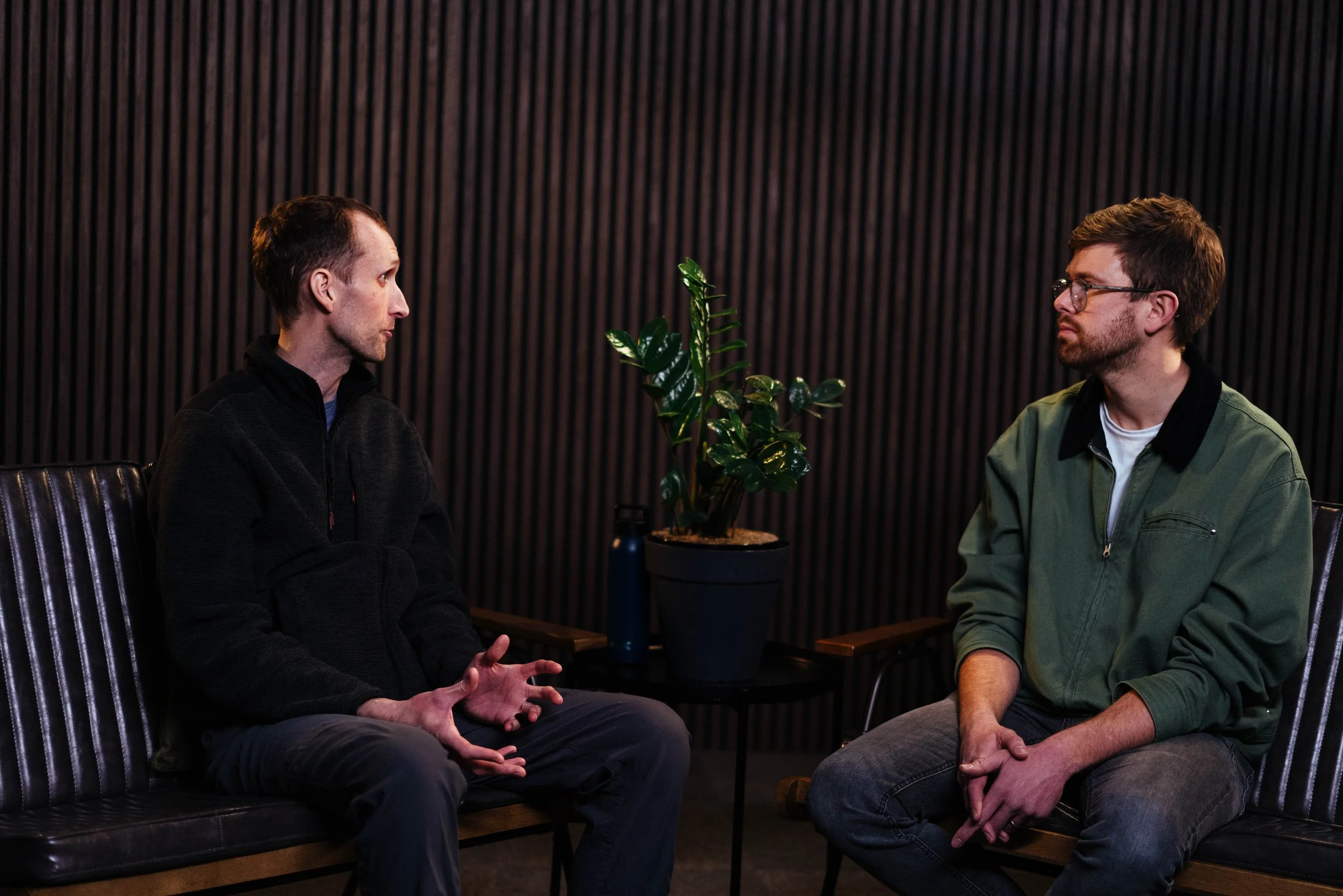 Two men sitting and talking in a room with dark wood paneling, a potted plant, and a small table with a water bottle behind them.