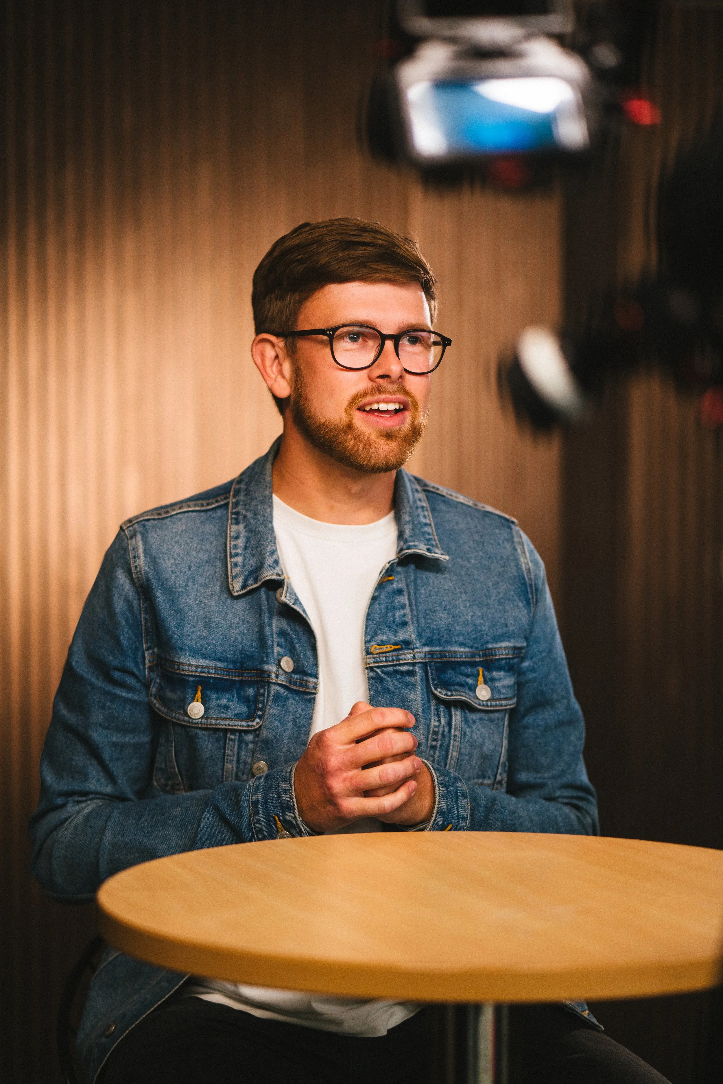 A young man with glasses, a beard, and a denim jacket sits at a round wooden table with hands clasped, speaking or giving a presentation, with a blurred background and a film crew camera in the foreground.